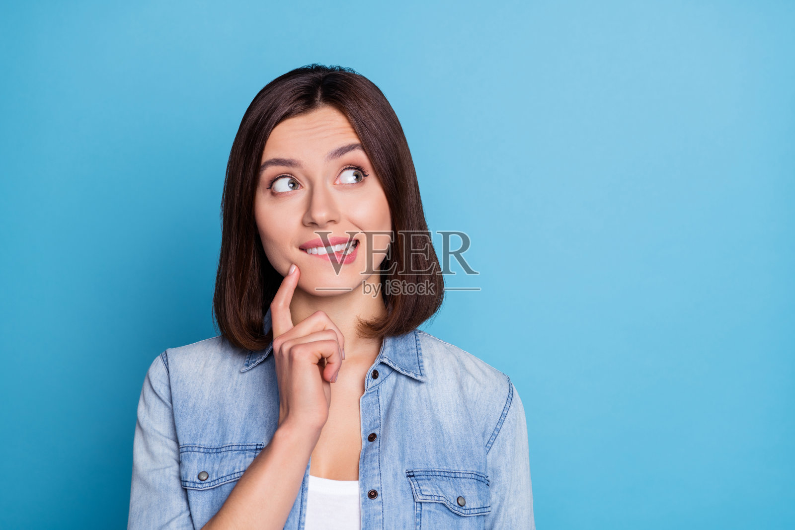 一位沉思的漂亮女士穿着牛仔衬衫,咬着嘴唇,凝视着空旷的空间,背景为蓝色。照片摄影图片