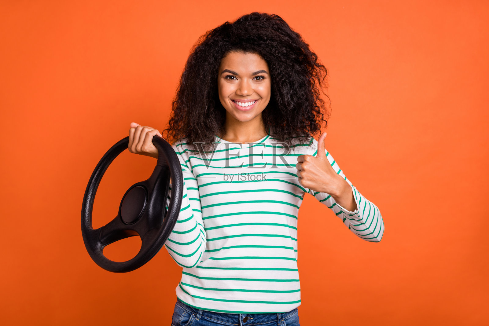 乐观的千禧一代女性,波浪卷发,开车时竖起大拇指,穿着衬衫,背景为橙色。照片摄影图片