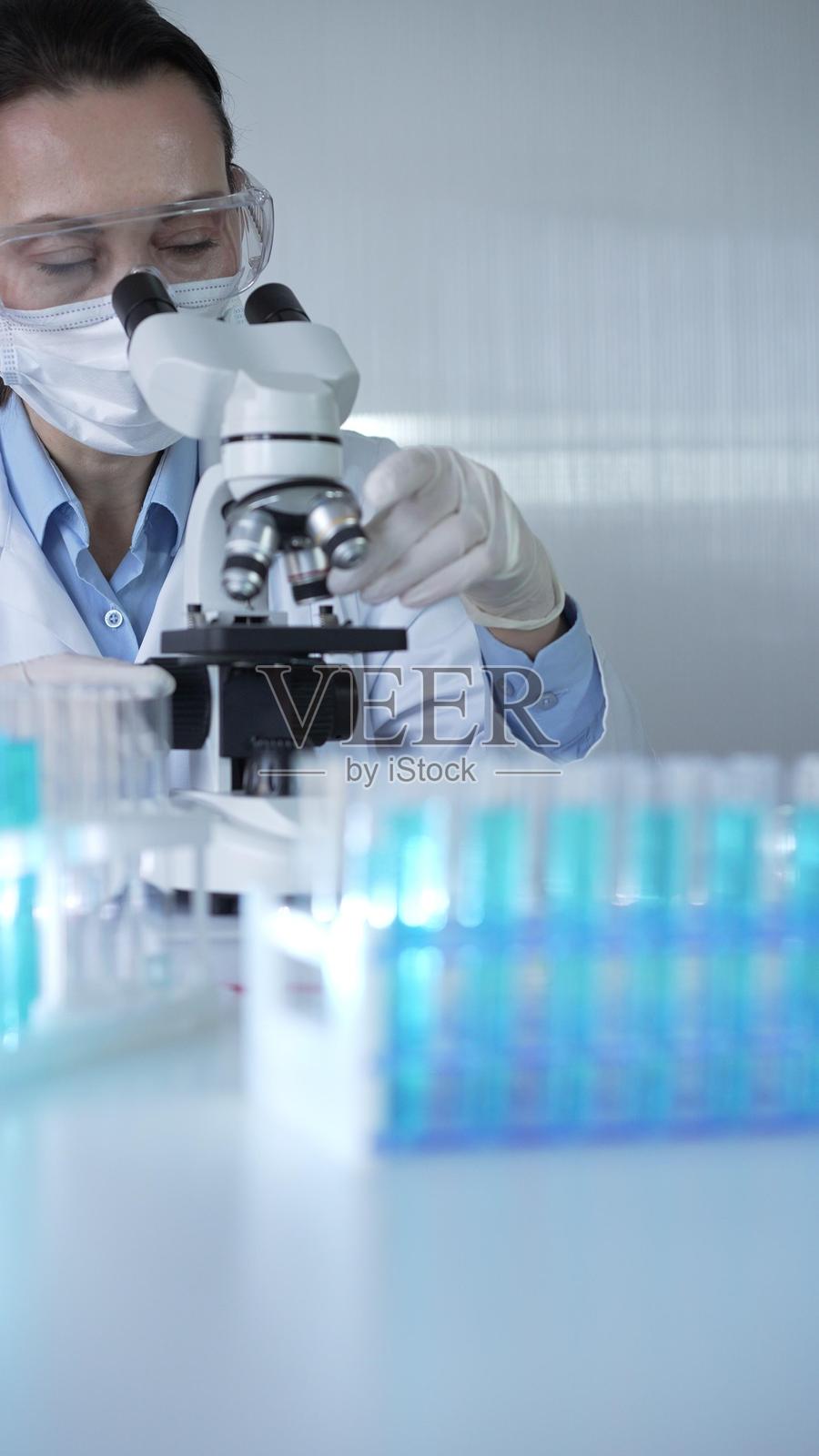 穿着实验室外套、戴着白色防护手套和眼镜的女科学家正在现代实验室中使用显微镜,肖像视图。低调照明照片。科学与医学概念。照片摄影图片