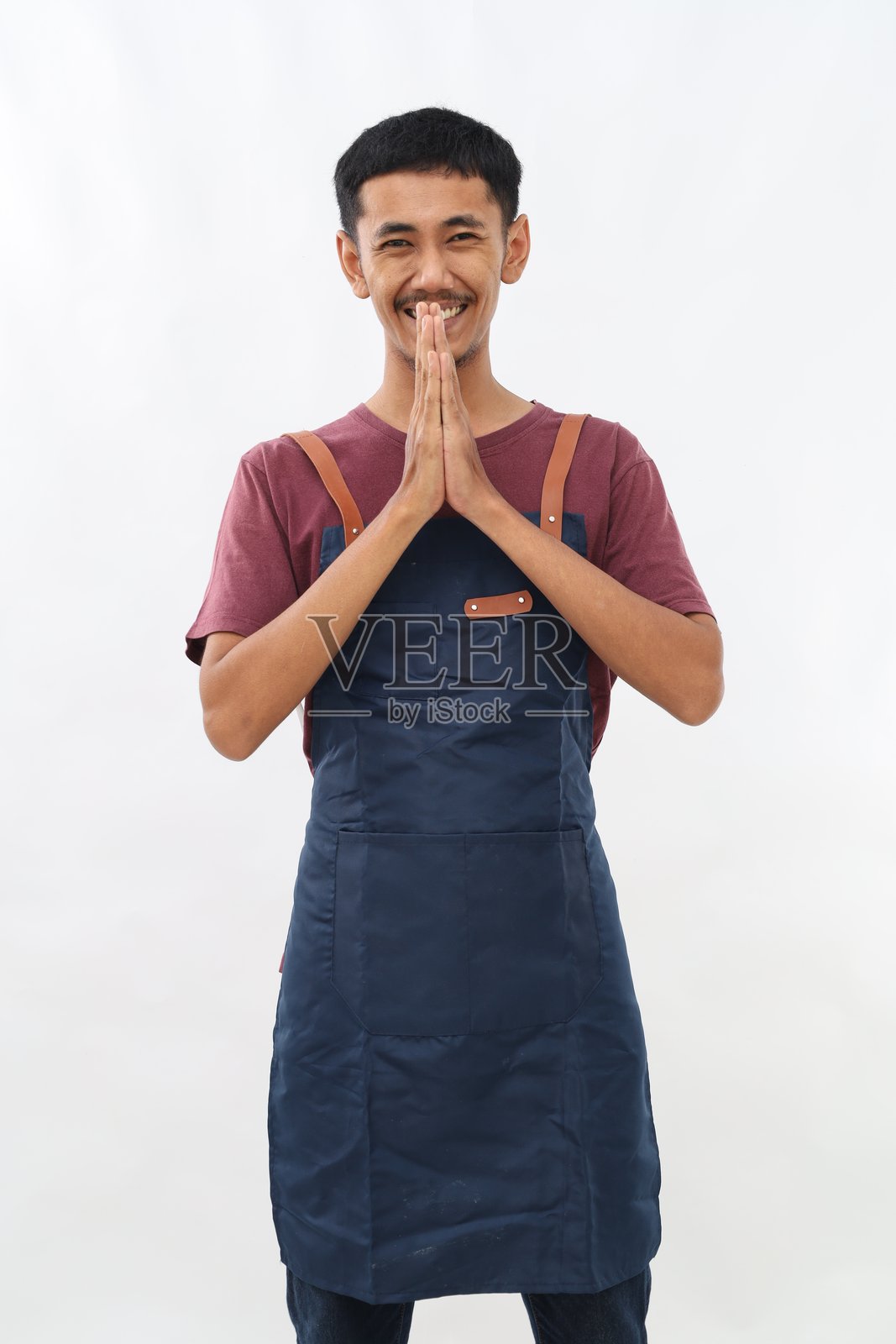 亚洲年轻男子穿着围裙,双手合十在白色背景下祈祷,面带微笑,自信地请求宽恕。照片摄影图片