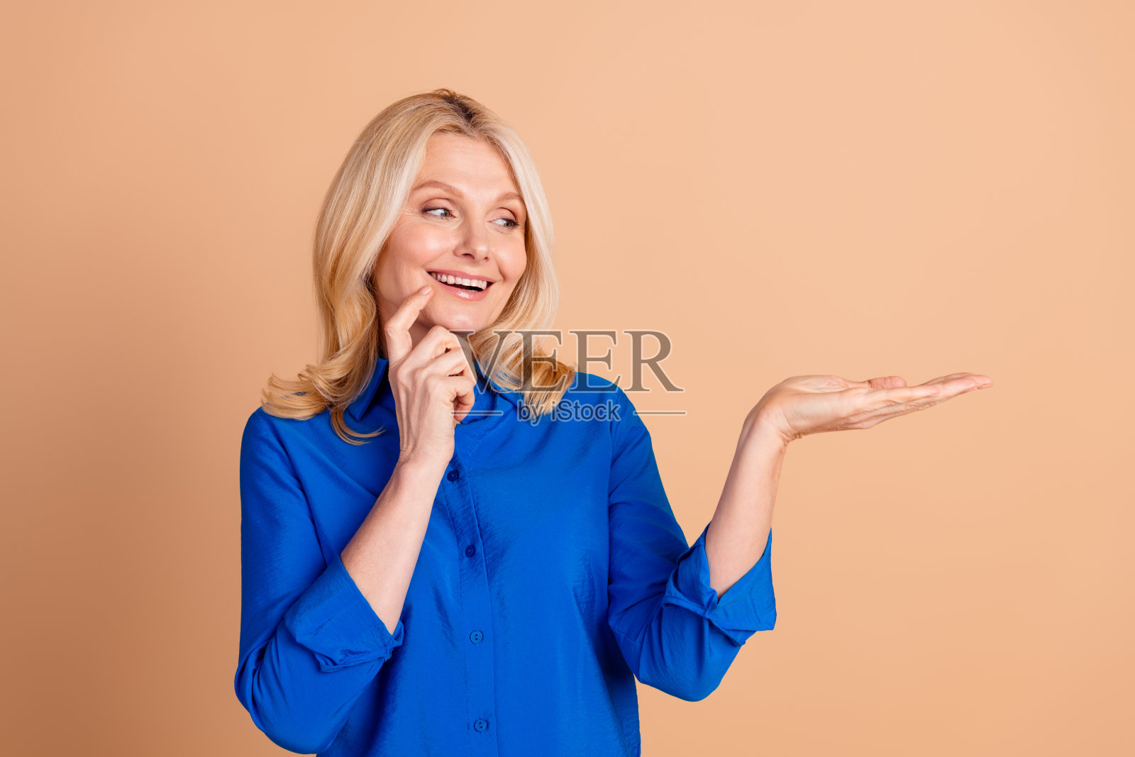 穿着蓝色衬衫的时尚成熟的女性,一只手做出手势,面带灿烂笑容,背景为米色。照片摄影图片