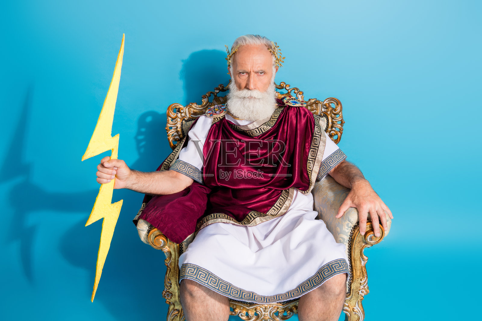 强壮的成熟男性坐在金色王座上,手握闪电,身穿古罗马皇帝长袍,背景为蓝色。照片摄影图片