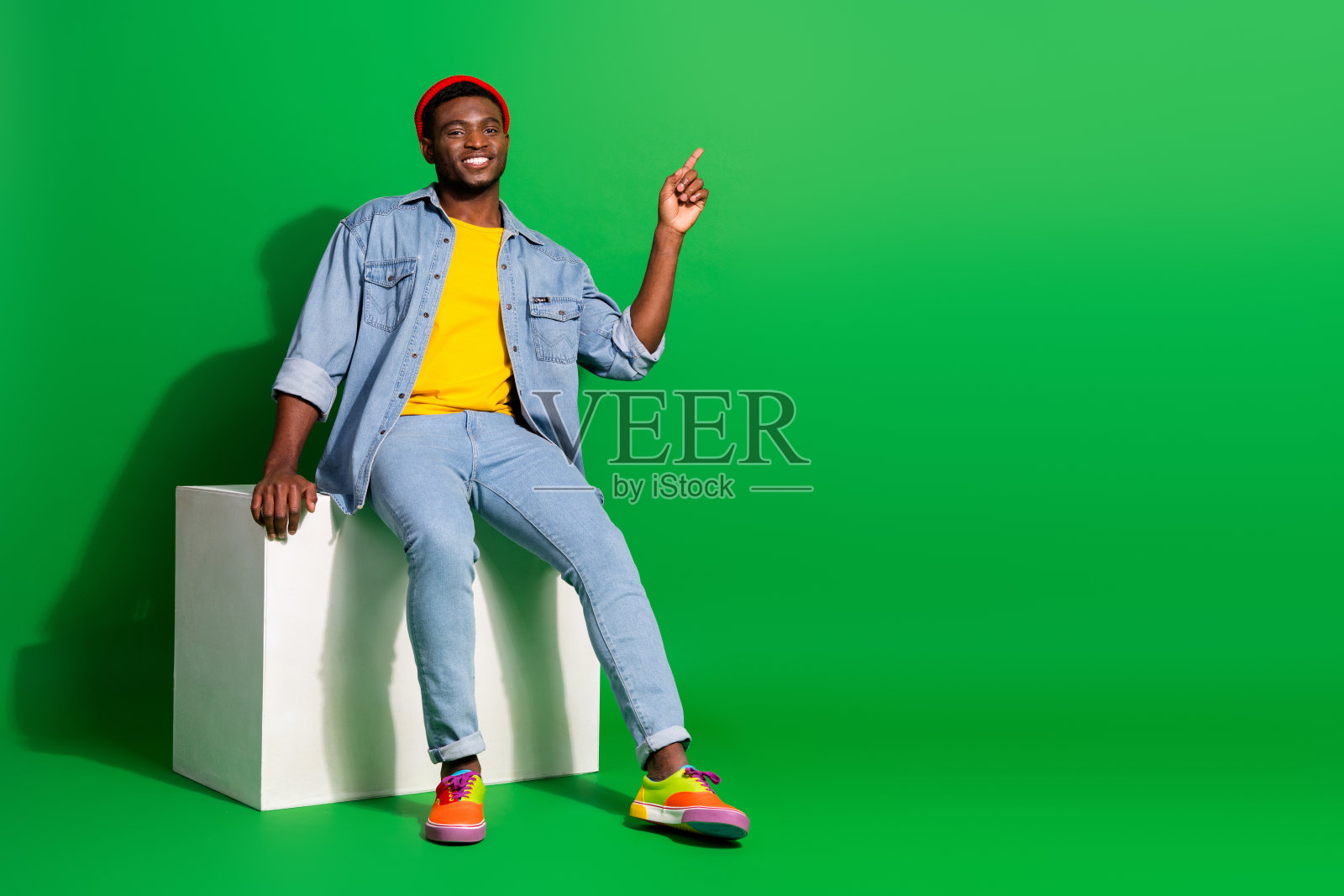 年轻时尚的男性坐在绿色背景前,穿着牛仔裤和色彩鲜艳的鞋子,表现出快乐和热情。照片摄影图片