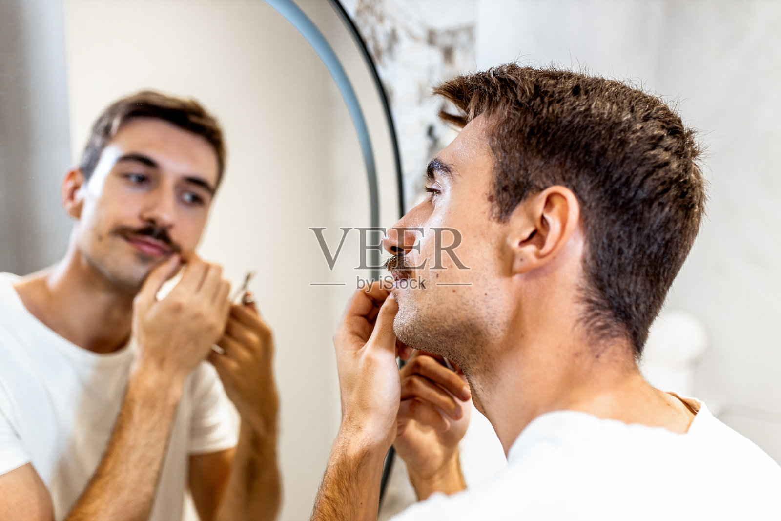 年轻男子在浴室镜子前用剪刀修剪胡须。照片摄影图片