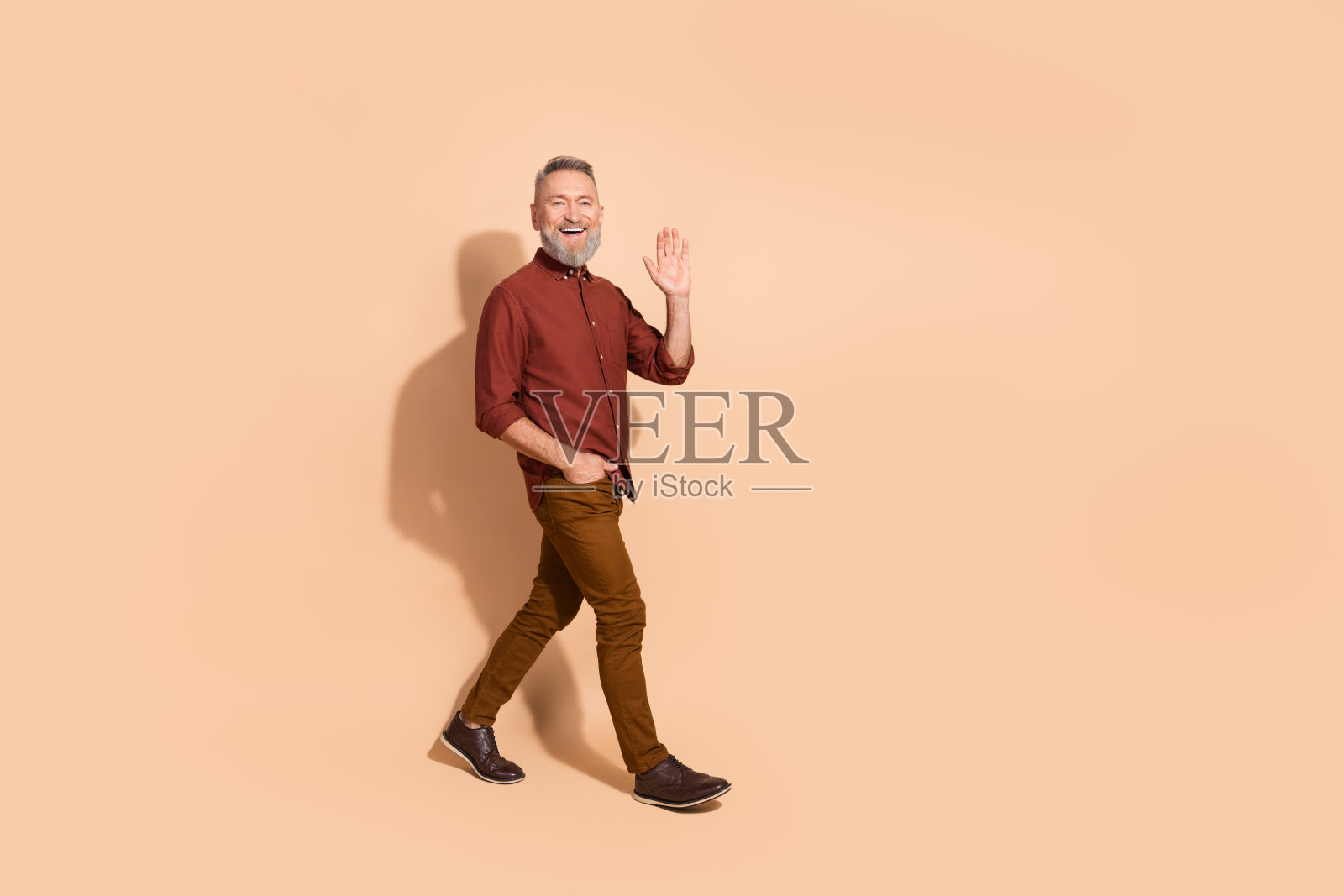 穿着时尚休闲服的成熟帅气男士,微笑自信地挥手走在米色背景前。照片摄影图片