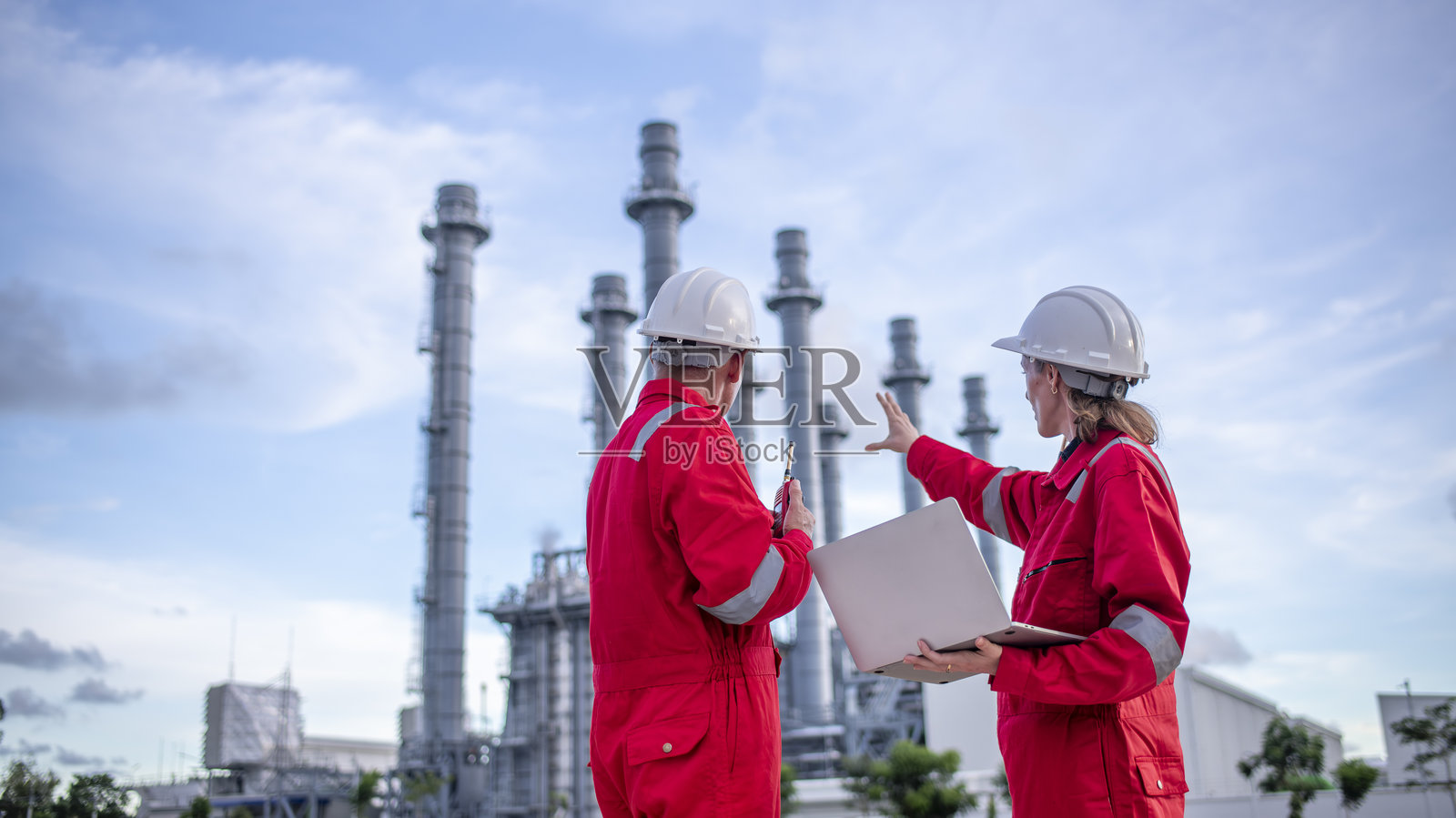 工程师们穿着安全装备,包括安全帽,正在检查一张大型蓝图平板,站在一个需要高安全标准的工业设施,如天然气或石油炼油厂,进行工作。照片摄影图片