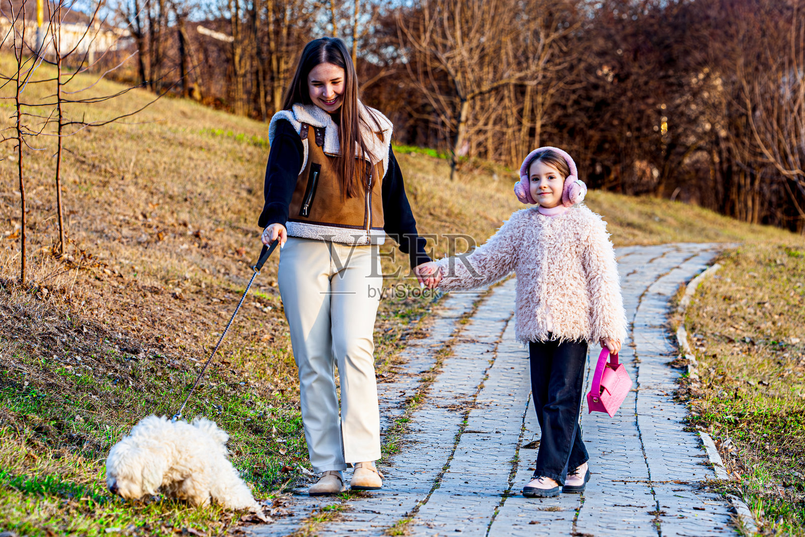 两个可爱的小女孩在绿色公园里牵着一只非常毛茸茸的白狗,脸上洋溢着巨大的快乐。照片摄影图片