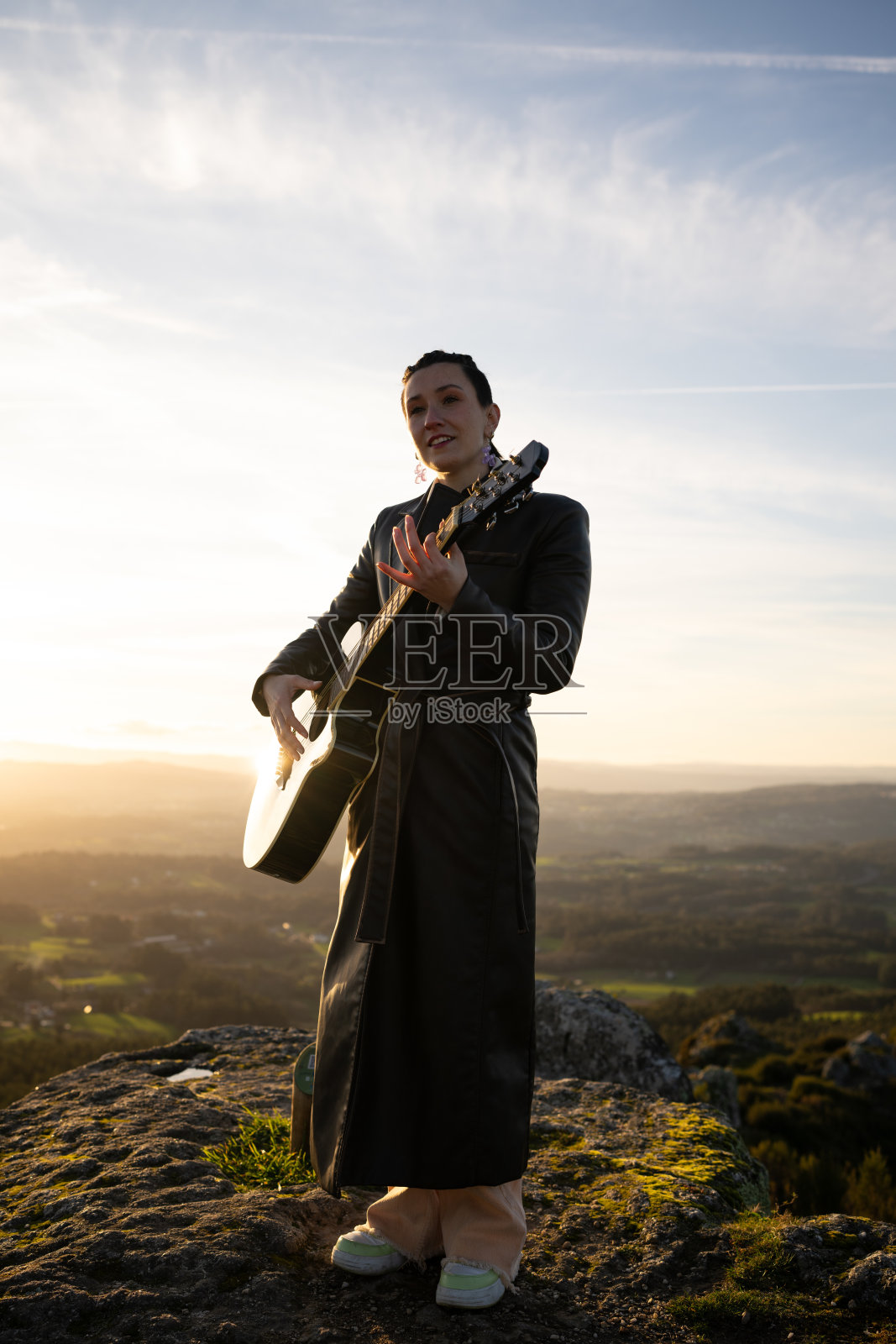 女性吉他手在岩石山峰上演奏原声音乐,剪影映衬在绚丽的日落天空和壮丽的自然背景中。照片摄影图片