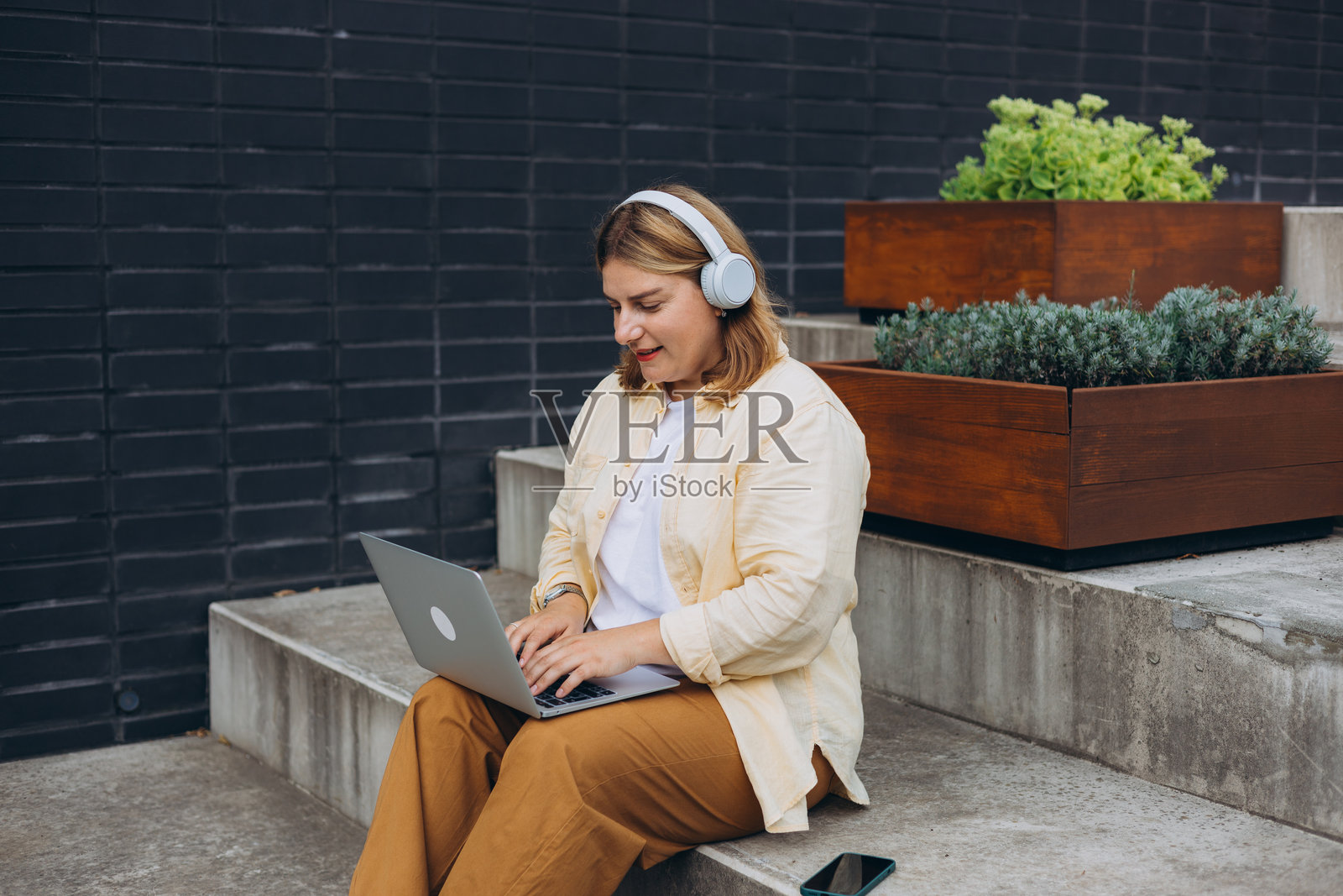 戴着耳机的快乐30岁女性在笔记本电脑上做生意并微笑。年轻女性在户外享受她的工作。远程工作,自由职业。漂亮的女性在城市街道的台阶上工作。照片摄影图片