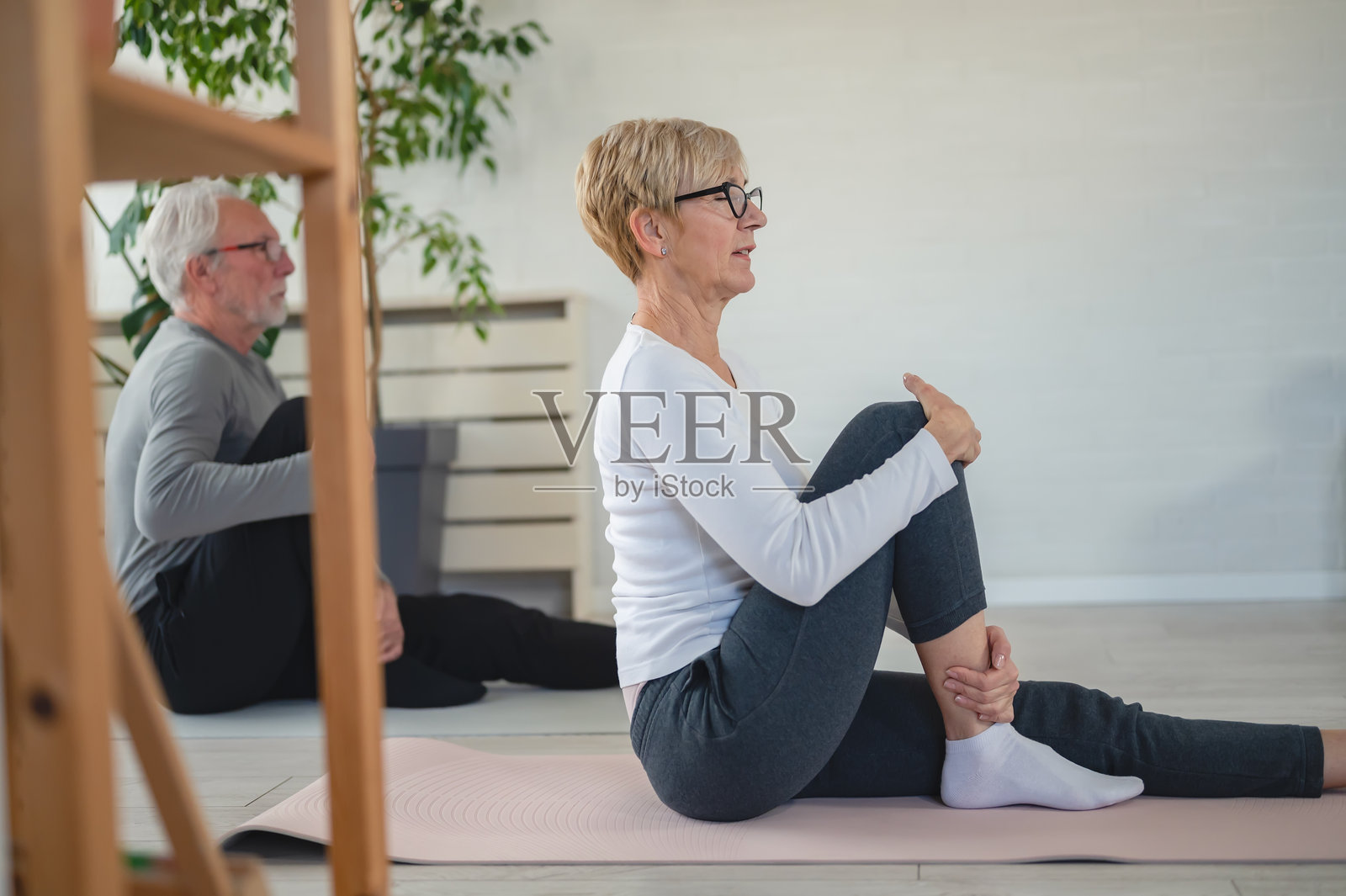 老年夫妇在家练习瑜伽,坐在垫子上进行坐姿脊柱扭转拉伸。他们身处一个明亮的客厅,阳光透过窗户洒入,周围有室内植物。照片摄影图片