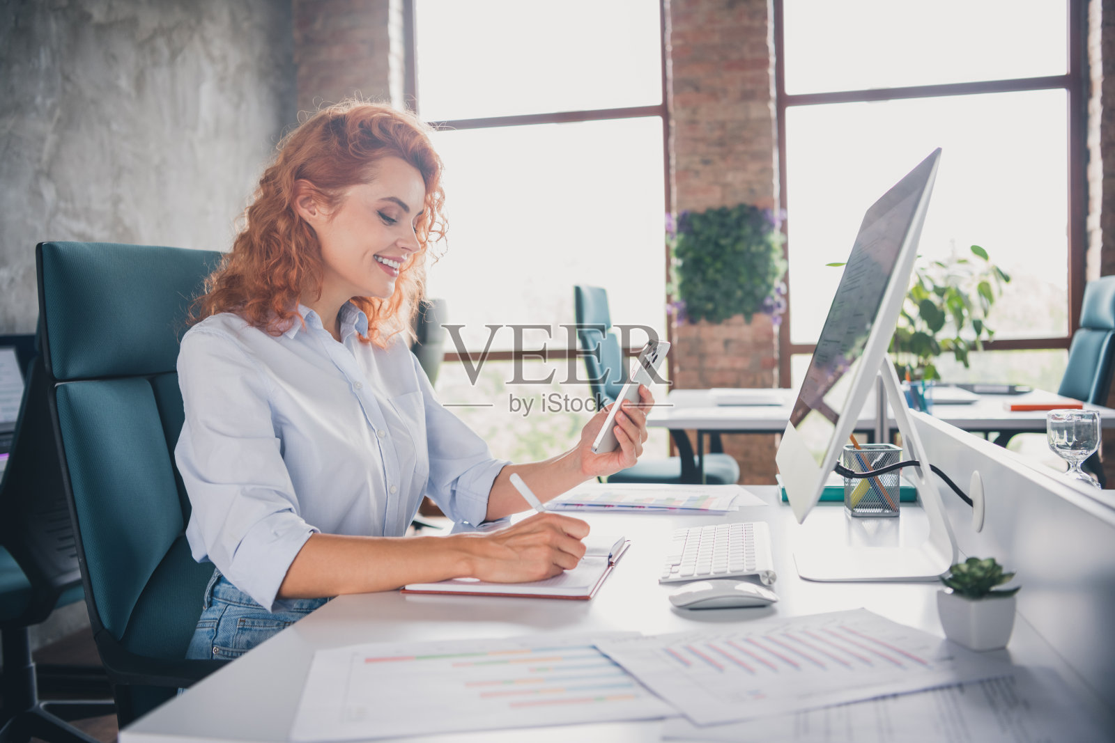 成功的年轻女商人手持手机,在现代阁楼风格的室内办公室桌上写笔记本。照片摄影图片