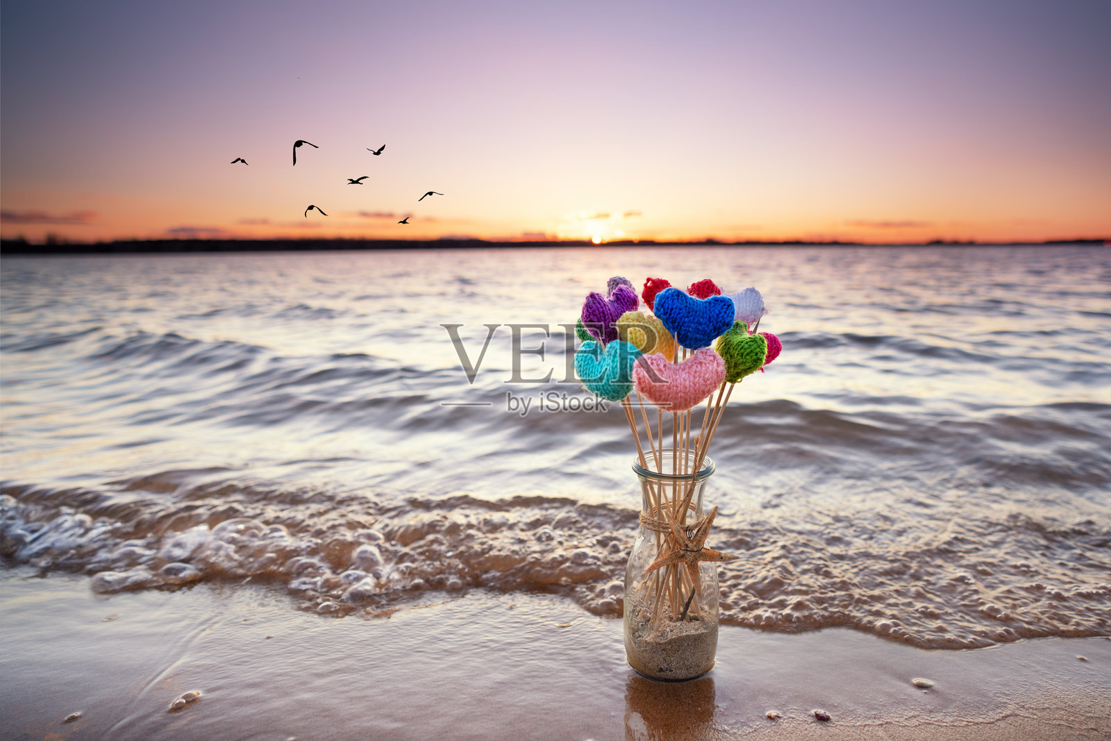 沙滩上夏日日落时,沙子里放着一个装满编织彩色心形的花瓶照片摄影图片