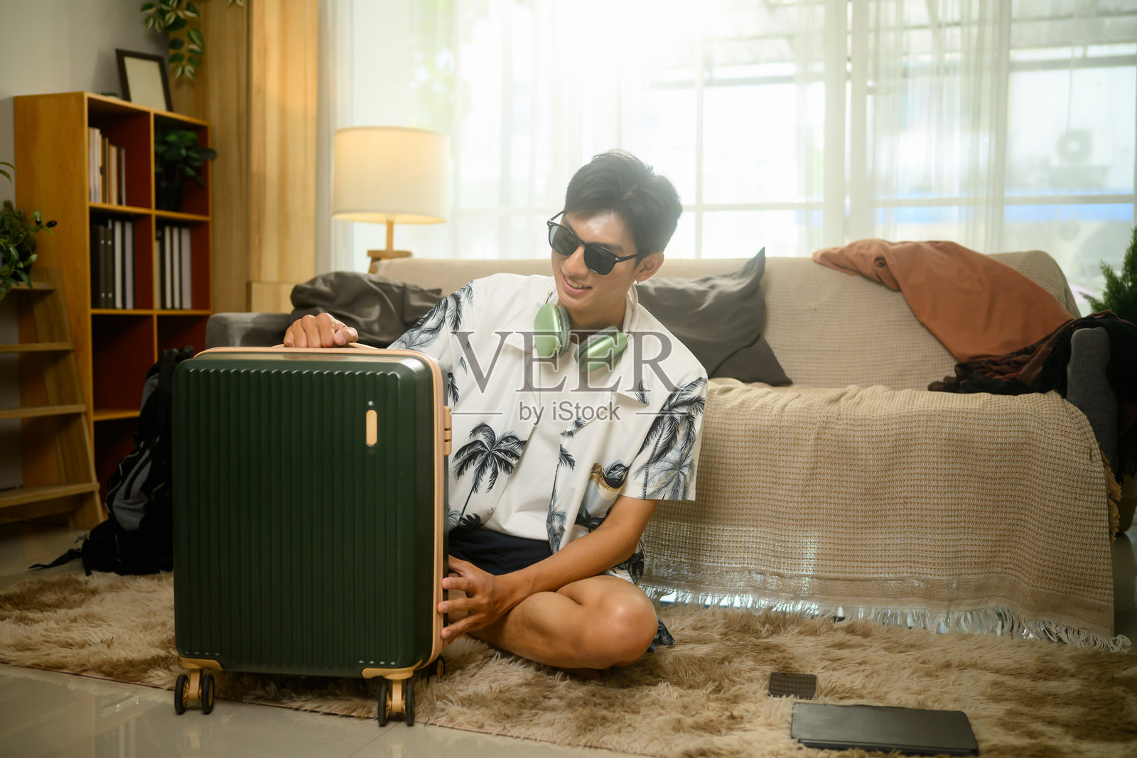 兴奋的旅行者穿着热带衬衫和太阳镜,坐在地板上与行李箱一起,准备他的夏季旅行。照片摄影图片