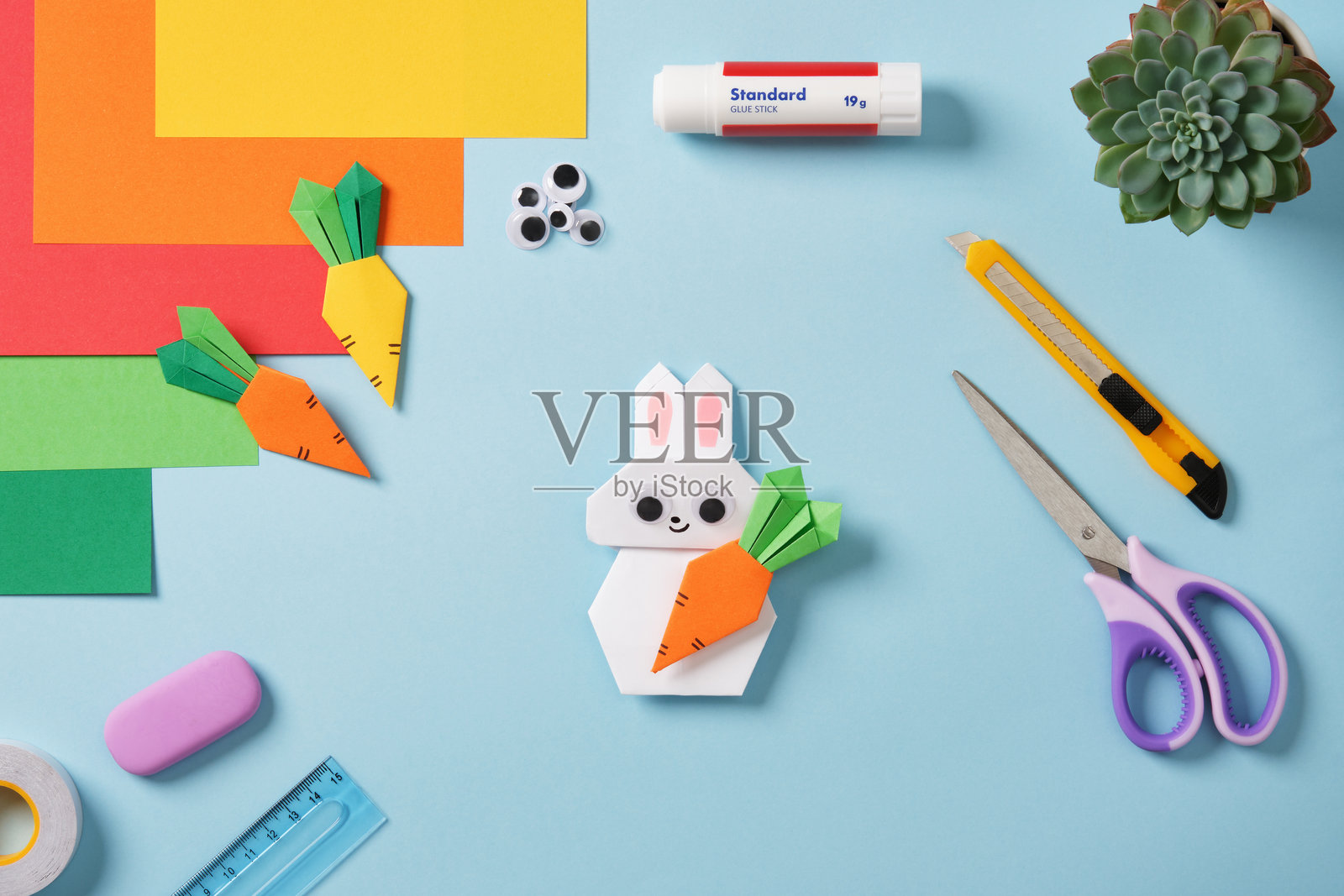 白色手工折纸兔子手持胡萝卜,还有其他纸胡萝卜、创意工具和材料:剪刀、刀具、胶水、尺子、橡皮擦。照片摄影图片