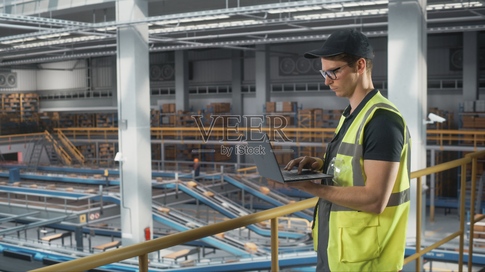 物流运输仓库设施:男性员工使用笔记本电脑监控设施的自动化操作。传送带正在装载在线购物订单的产品箱,以便送达客户。照片摄影图片