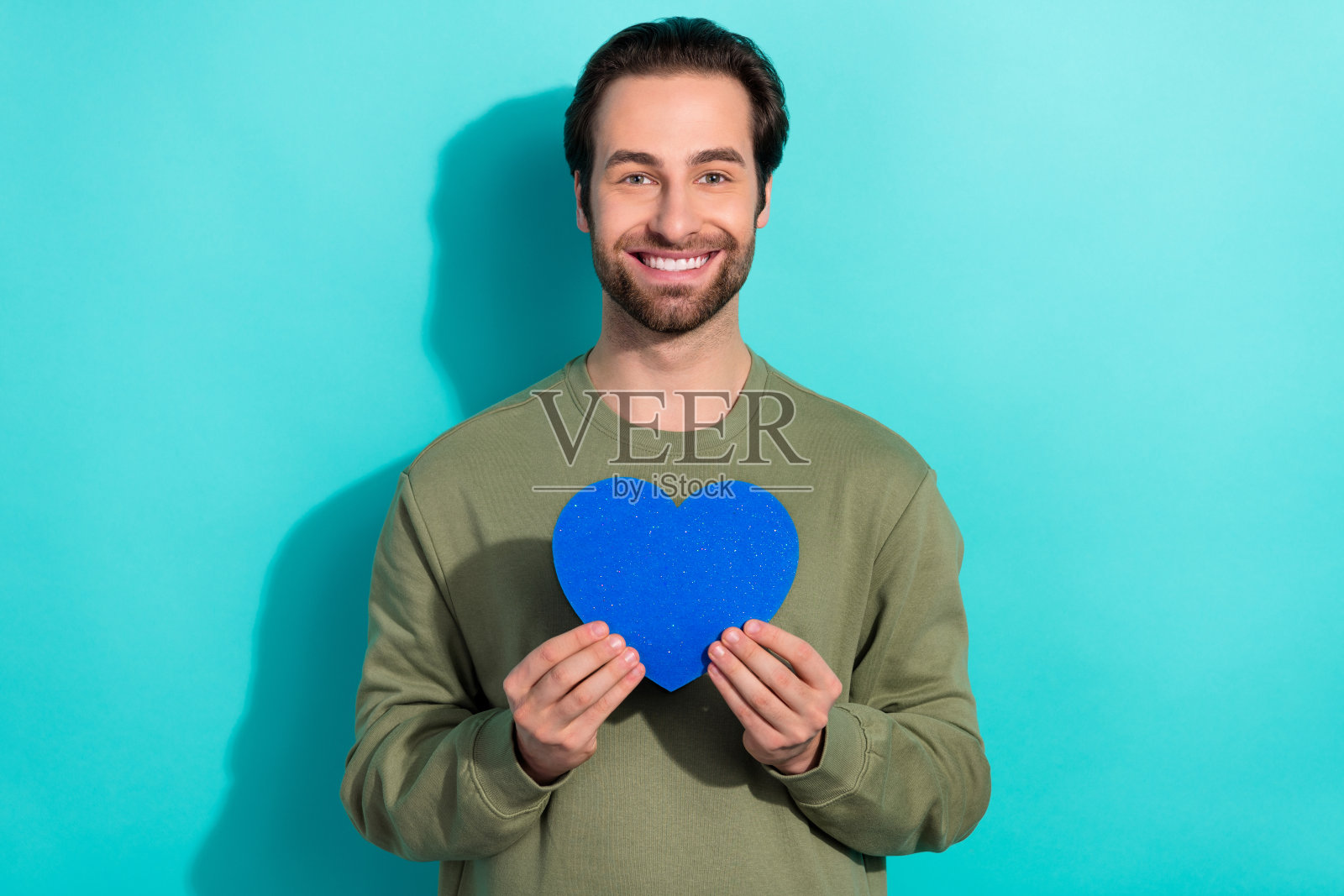 可爱的胡子千禧男孩穿着卡其色卫衣,手持心形物品,背景为青绿色。照片摄影图片
