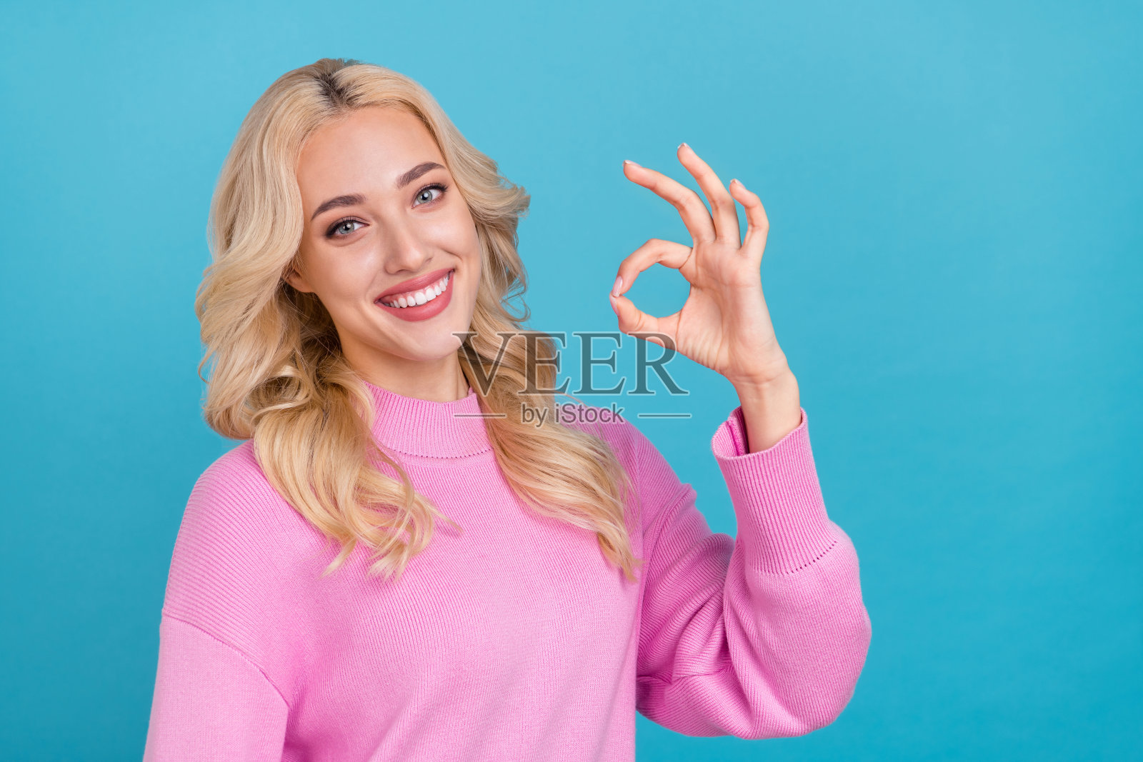 迷人的快乐波浪卷发女孩在明亮的蓝色背景下做出“好”的手势。照片摄影图片