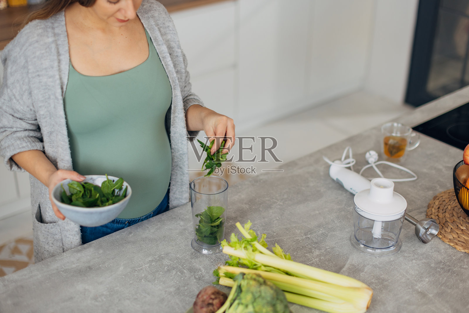 一位孕妇在厨房为午餐准备有机沙拉。她将绿色蔬菜叶放入搅拌机中。照片摄影图片