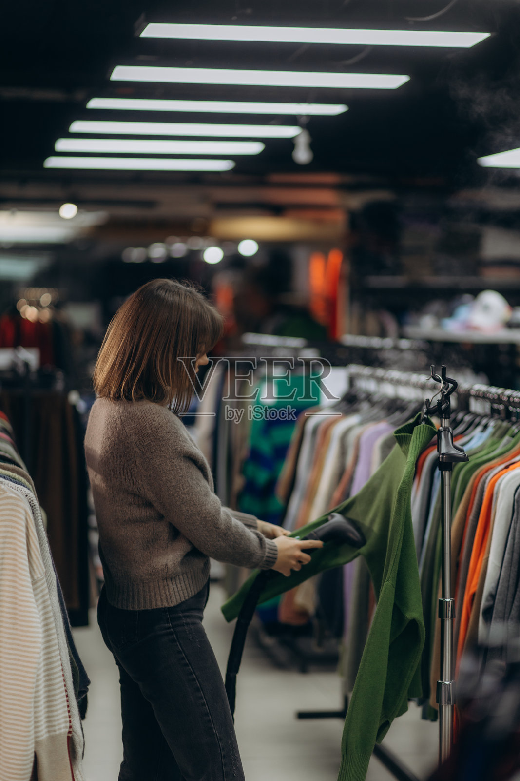 在时尚室内灯光下,穿着绿色毛衣的人在服装店里蒸熨衣物照片摄影图片