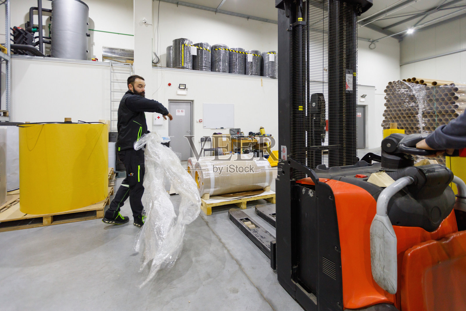 男子在配送仓库中拿着塑料包装膜,同事正在使用叉车。照片摄影图片