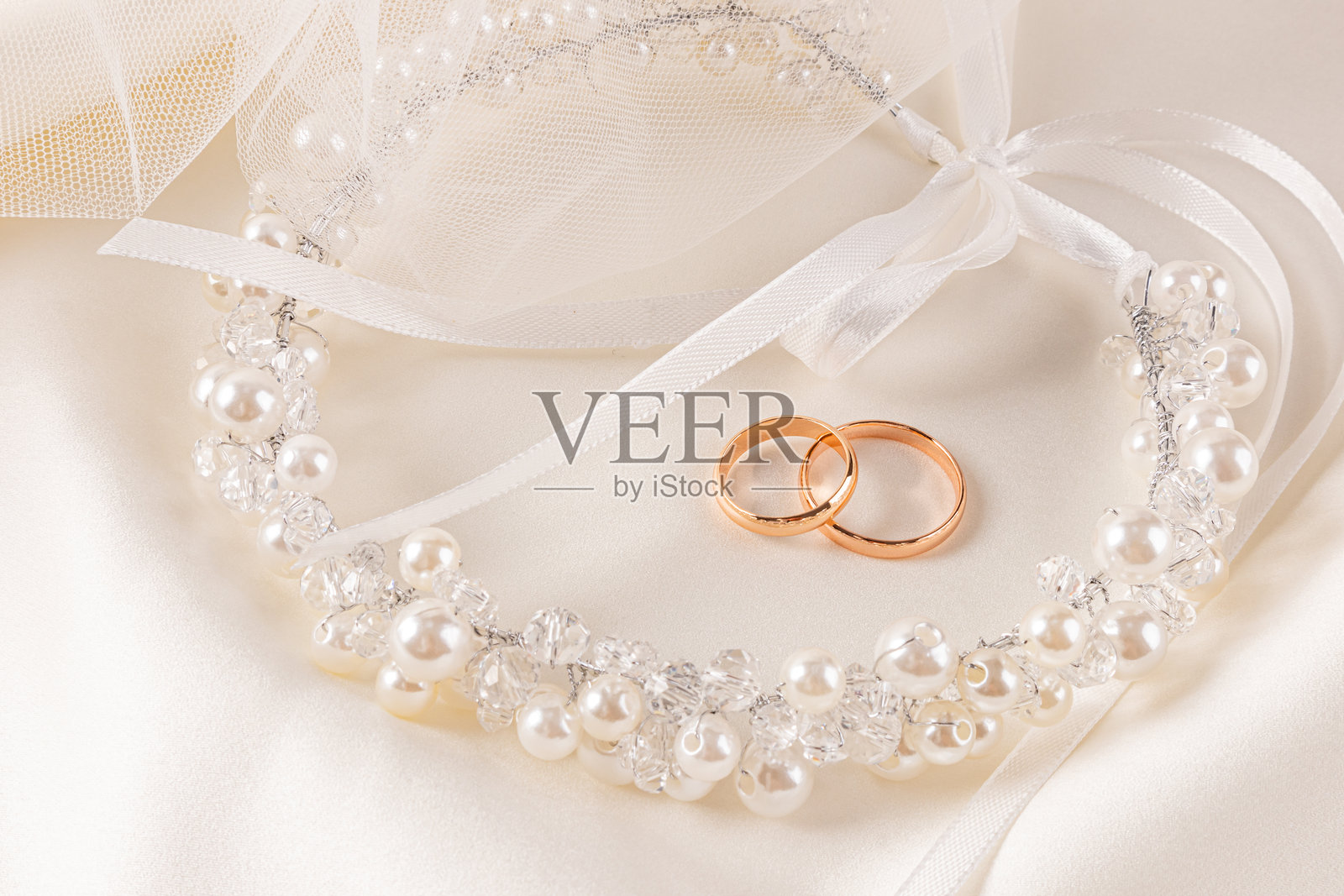 两枚金色婚戒静静地躺在米色缎面背景上,背景中有新娘发型的时尚装饰,配有珍珠花环和丝带。明信片。照片摄影图片