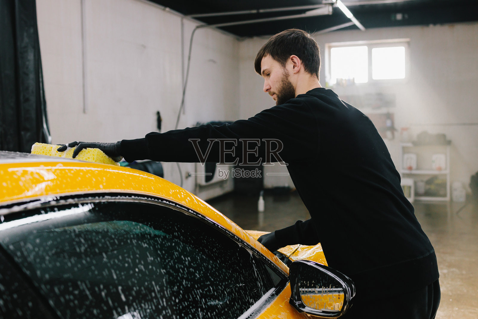 在车库里用黄色海绵和泡沫手洗跑车车身。洗车工正在进行手动泡沫清洗,提供汽车美容服务。照片摄影图片