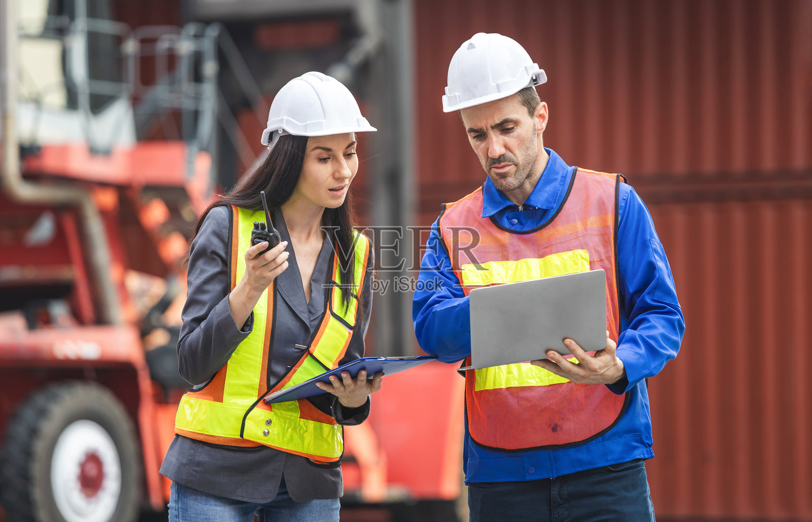 穿着安全装备的女性主管使用夹板检查清单,工头工人戴着安全帽手持笔记本电脑,控制装载货物集装箱。照片摄影图片