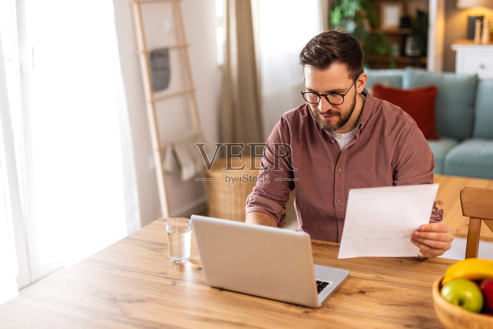 年轻男子在家中用笔记本电脑工作时微笑。舒适的客厅里有书架、温暖的灯光、室内植物和一碗新鲜水果,营造出一个平衡的工作环境。照片摄影图片