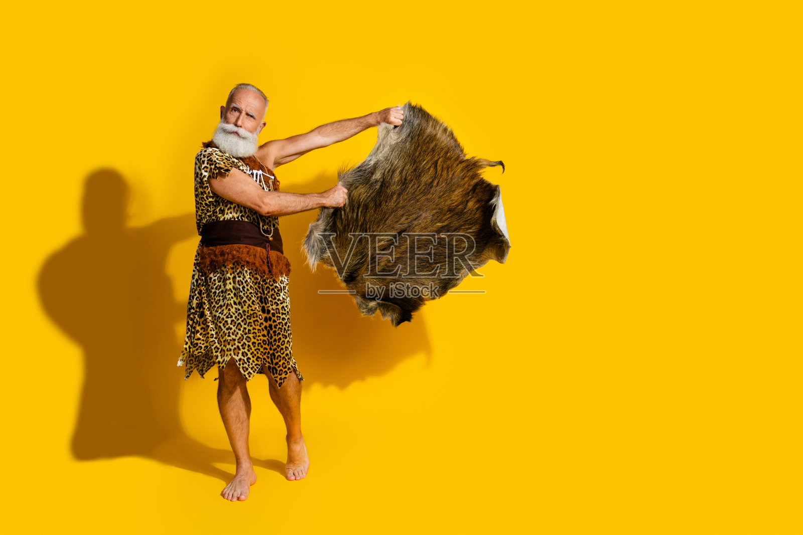 全身照片,古老的穴居人穿着豹纹衣服,手持动物皮毛,空旷的空间,背景为黄色。照片摄影图片