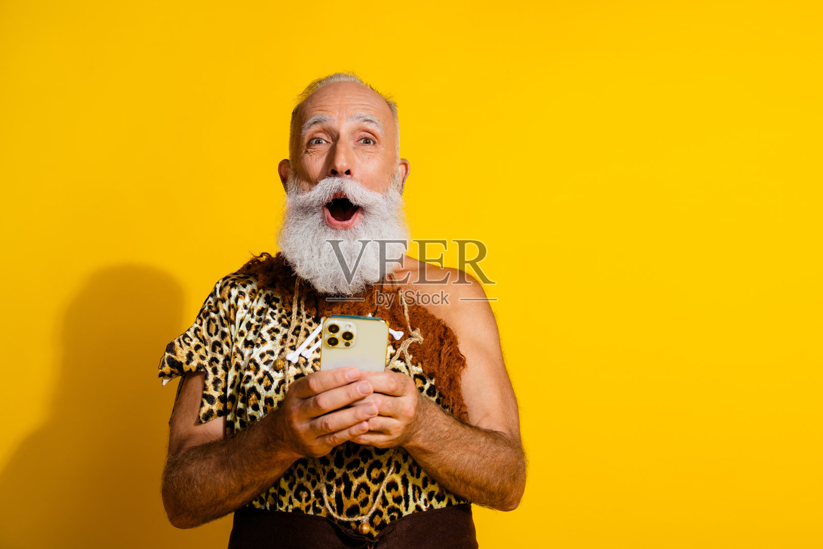 原始穴居人穿着豹纹服装,手持手机,站在黄色背景前,空旷的空间。照片摄影图片