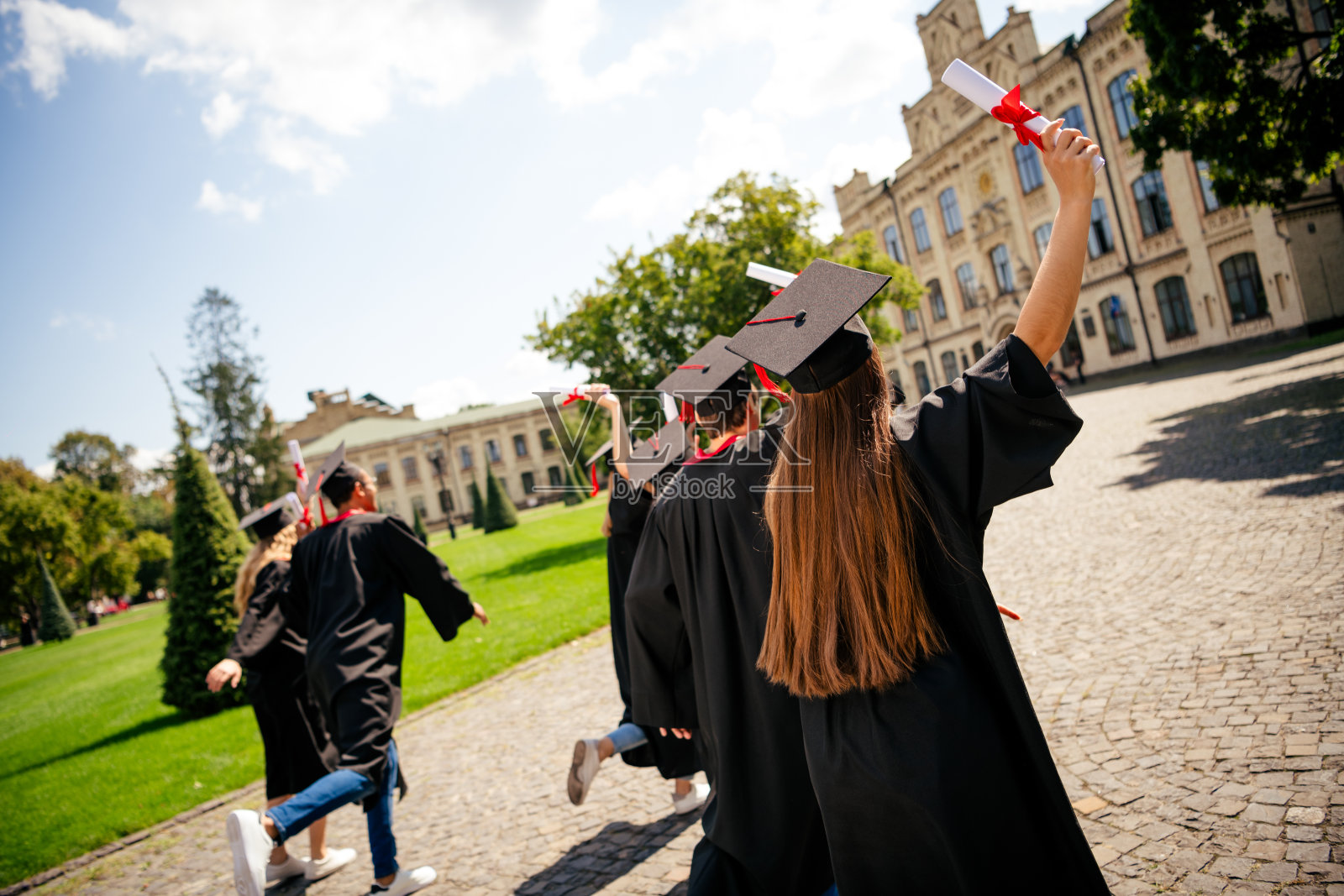 一群学生在户外大学建筑前,身穿毕业袍,手持文凭,背影奔跑,戴着学位帽。照片摄影图片