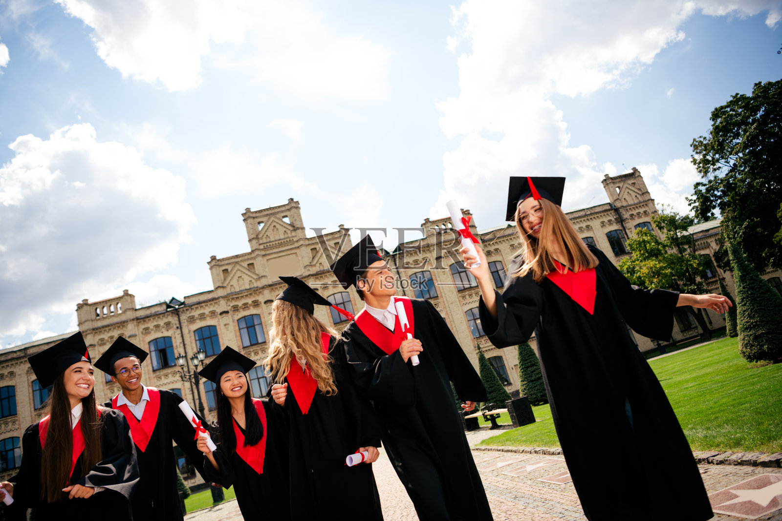 公司同学们在户外的大学建筑前,面带灿烂笑容,手持文凭,戴着学位帽,身穿毕业袍。照片摄影图片