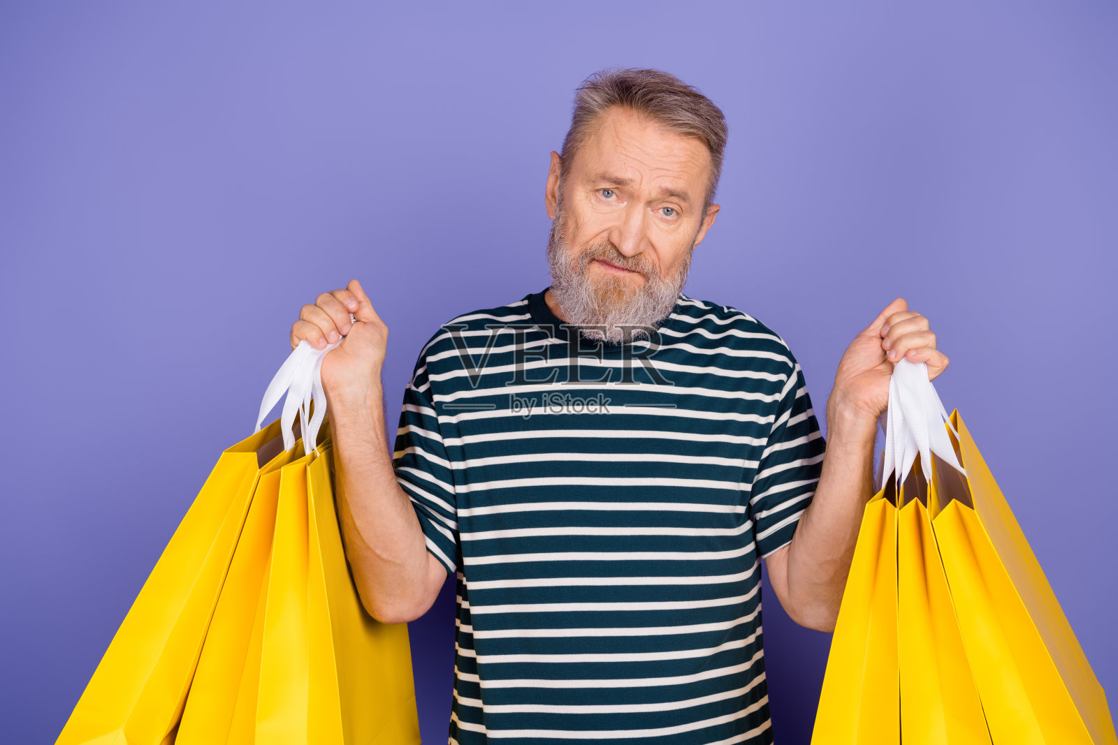 一位时尚的老年男子,留着胡须,手提鲜艳的黄色购物袋,背景为紫色。照片摄影图片