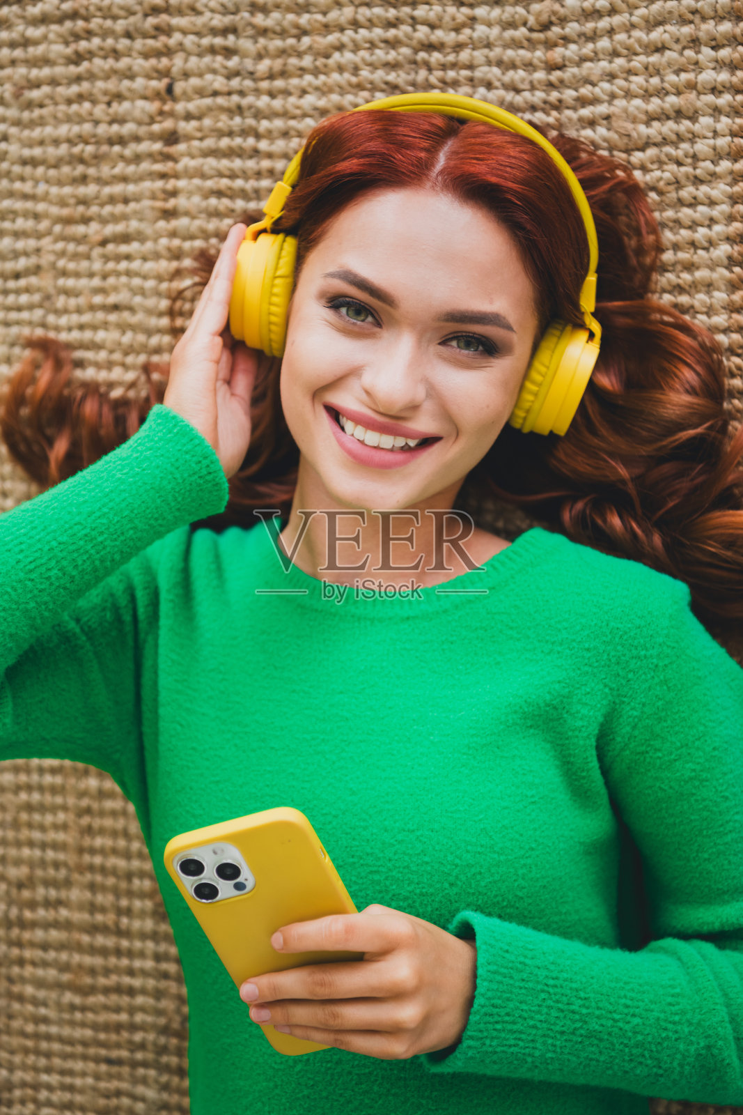 年轻漂亮的女子躺在地板上,戴着耳机,使用智能手机,穿着毛衣,享受周末的室内休闲时光。照片摄影图片