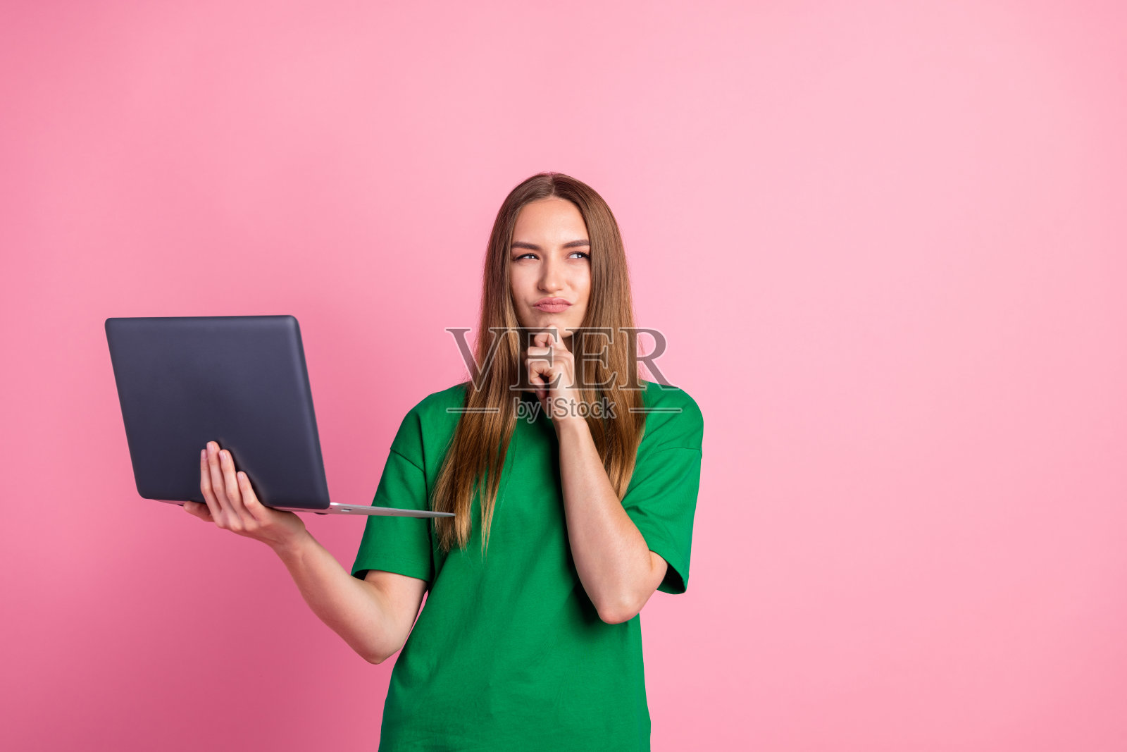 一位穿着休闲绿色衬衫的年轻女性,手持笔记本电脑,背景为粉色,展现出好奇心和现代生活方式。照片摄影图片