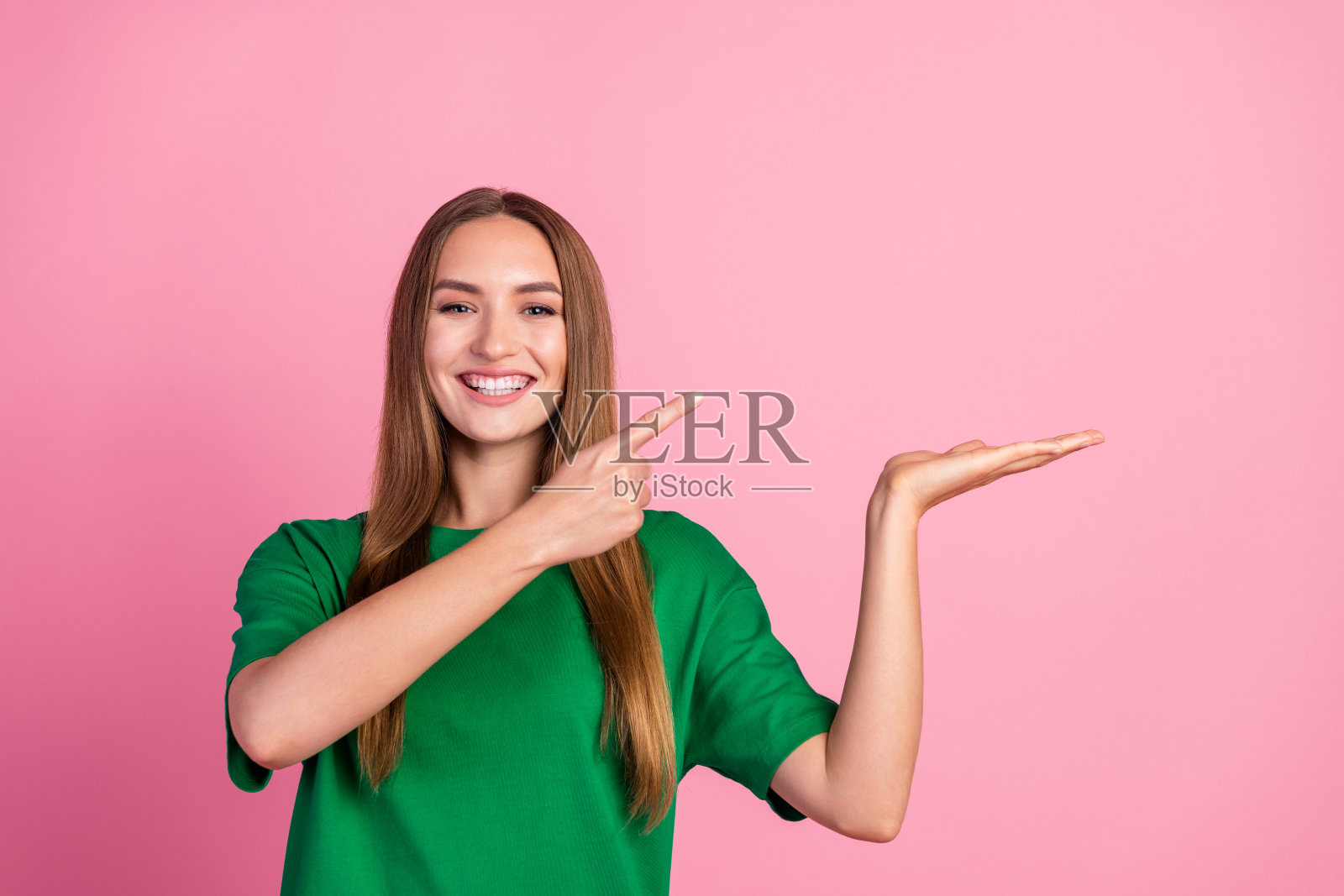 年轻女性在粉色背景上用手势表达,穿着绿色衬衫,开心地微笑,展现出愉快的活力。照片摄影图片