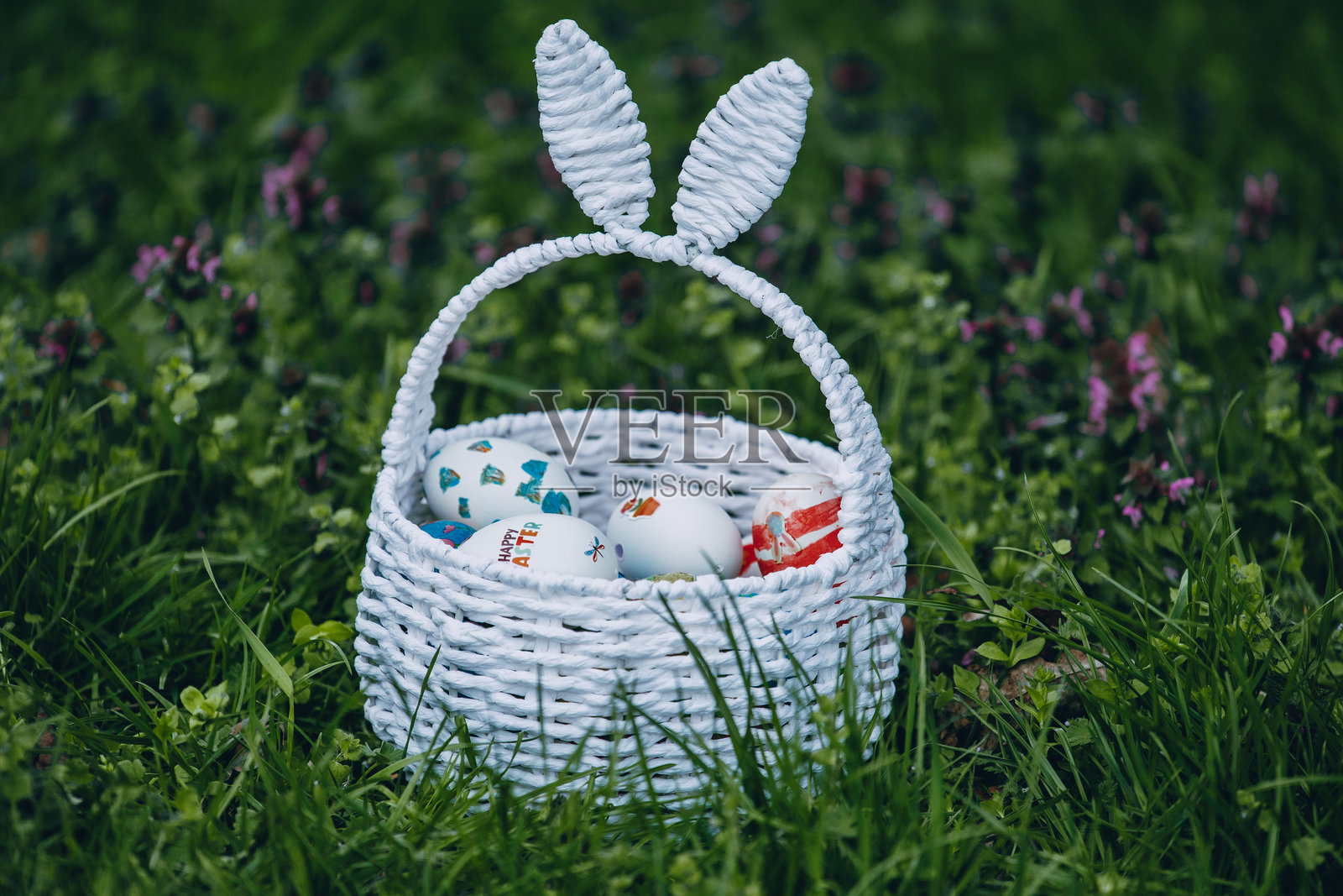 五彩缤纷的复活节彩蛋放在编织的篮子里,兔耳朵放在春季庆祝活动中的草地上。照片摄影图片