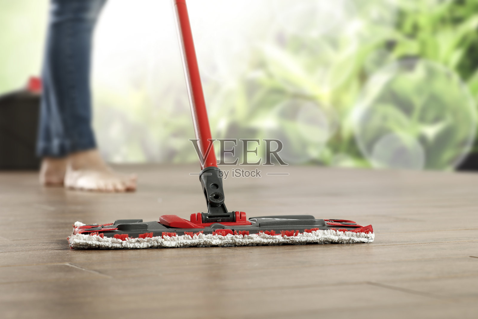 红色手柄的拖把配有矩形清洁垫,放在光滑的木地板上。赤脚和穿着牛仔裤的成年人的腿。正在家中清洁。照片摄影图片