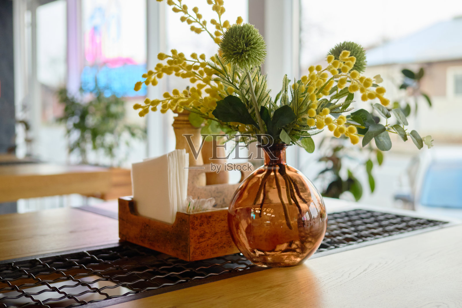 一个温馨宜人的咖啡馆展示了一张精美布置的桌子,上面摆放着一个装满鲜花的装饰花瓶,衬托出现代的氛围。自然光透过大窗户洒入室内。照片摄影图片