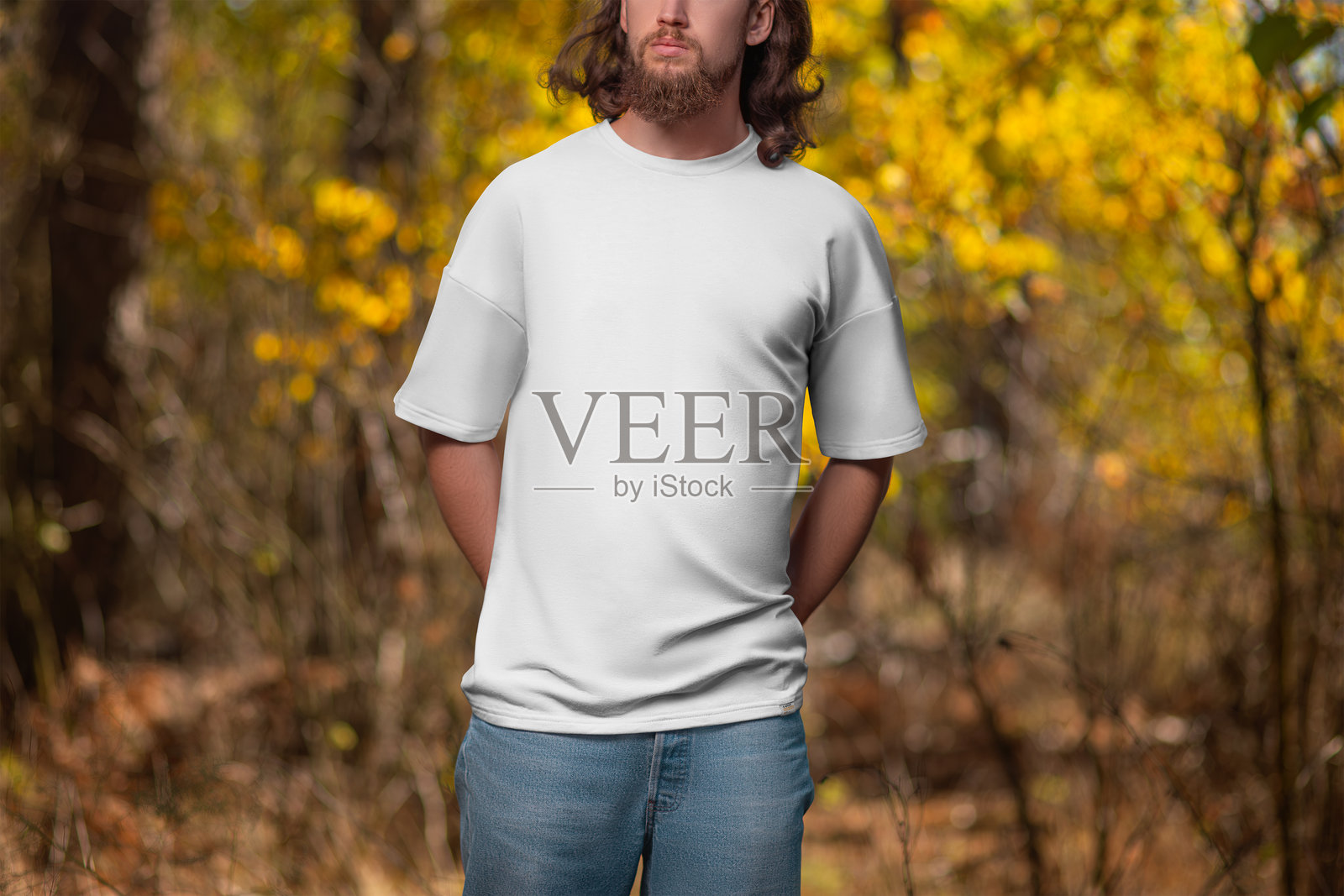 一位留着长发和胡子的男子穿着白色宽松T恤,背景是黄色的树木,正面视图。照片摄影图片