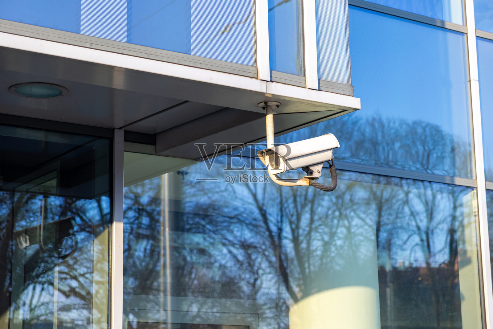现代玻璃建筑角落的安全监控摄像头,白天的光线中映射出树木和天空的倒影,城市建筑中的安全监控系统照片摄影图片