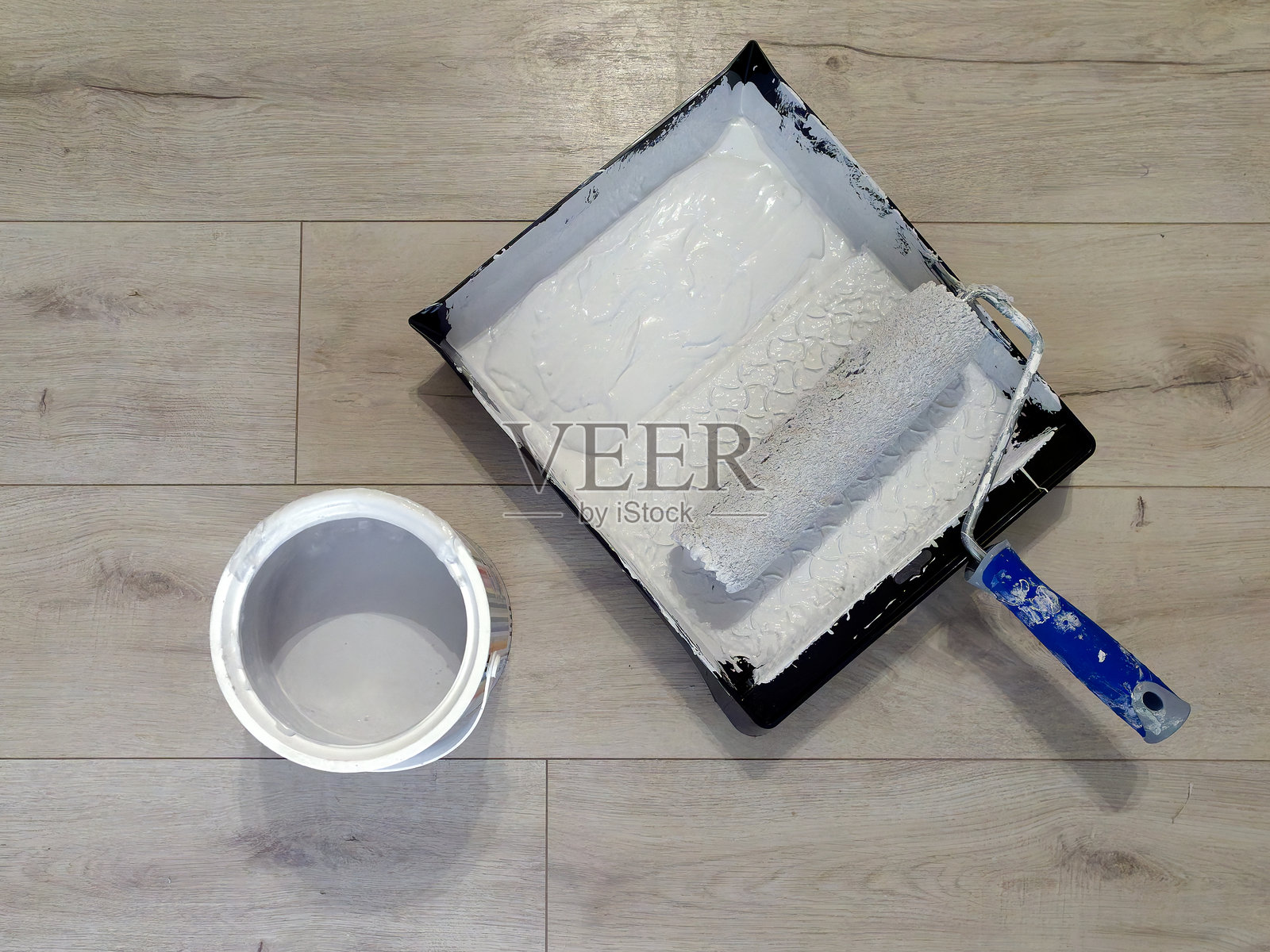 从上方看蓝色手柄的油漆滚筒放在灰色油漆托盘中,旁边有一罐灰色油漆,背景为自然木质纹理。照片摄影图片