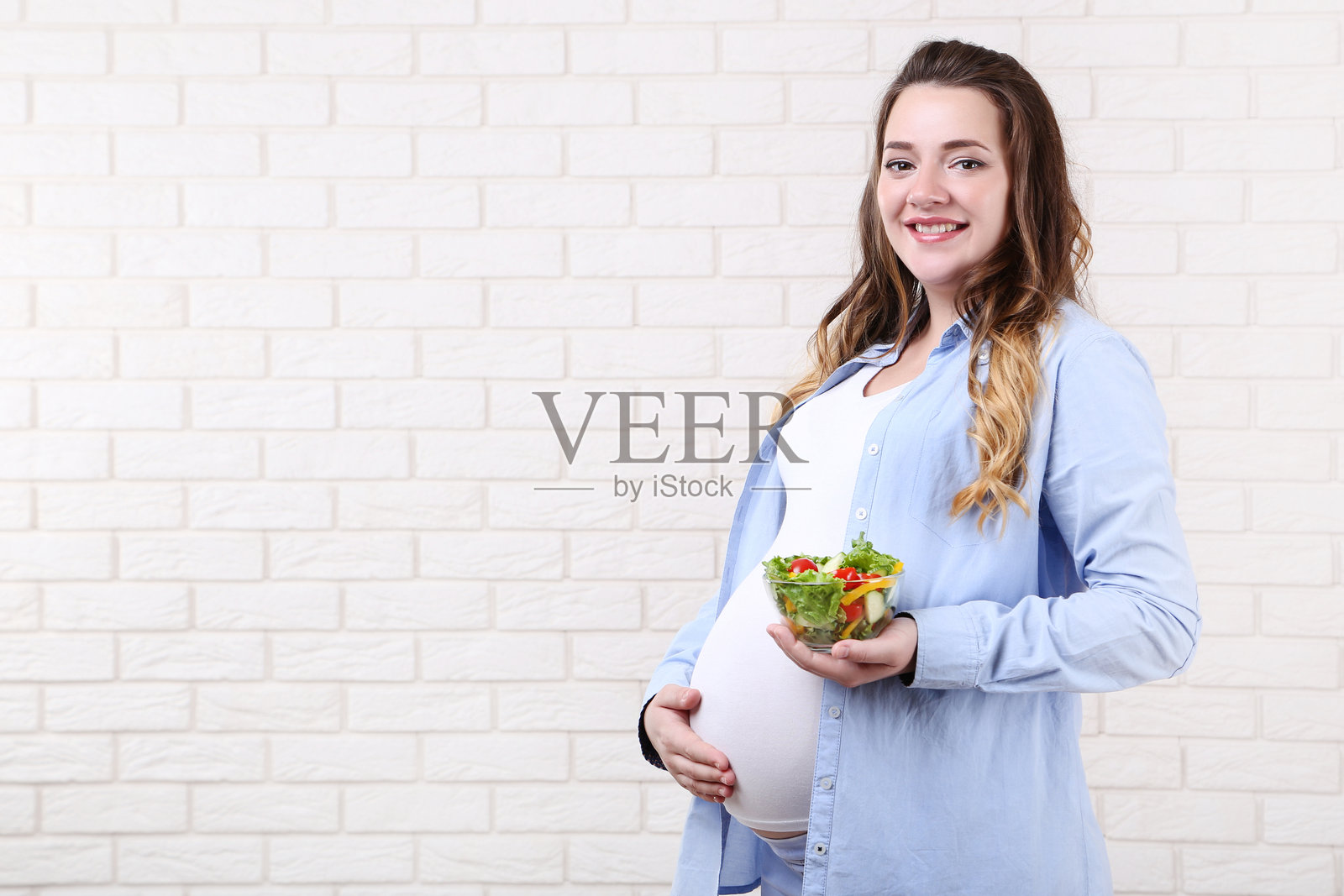 美丽的孕妇手捧沙拉碗,背景是砖墙照片摄影图片