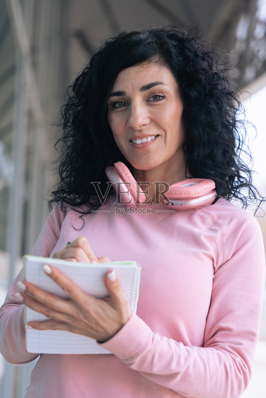 穿着粉色连帽衫的运动女性在户外戴着耳机写笔记本。照片摄影图片