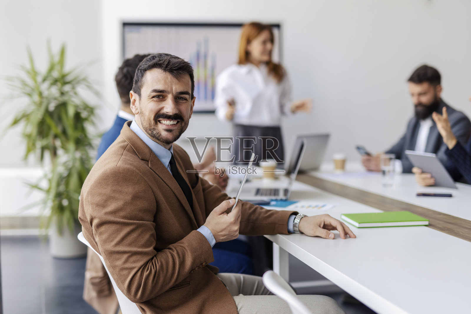 年轻商人在会议室中坐着,身后模糊的背景是他的团队成员,正在参加研讨会。照片摄影图片