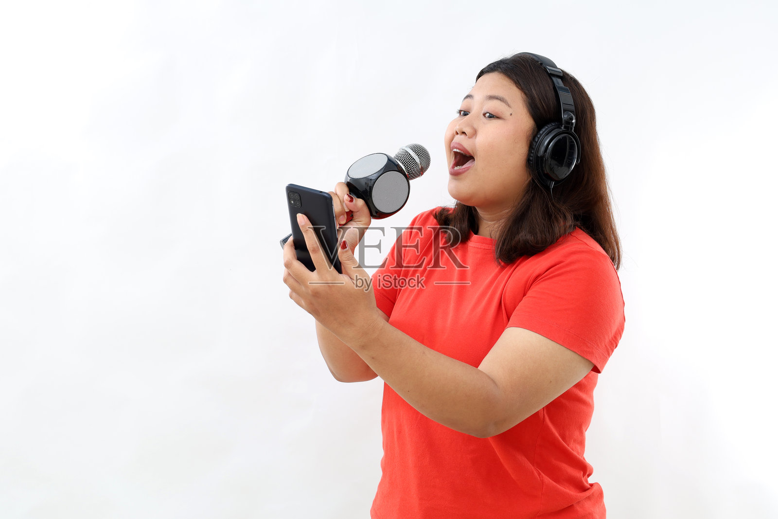 美丽的亚洲女性在白色墙壁前,穿着休闲T恤,戴着耳机听音乐,同时用麦克风唱歌,正看着手机。照片摄影图片