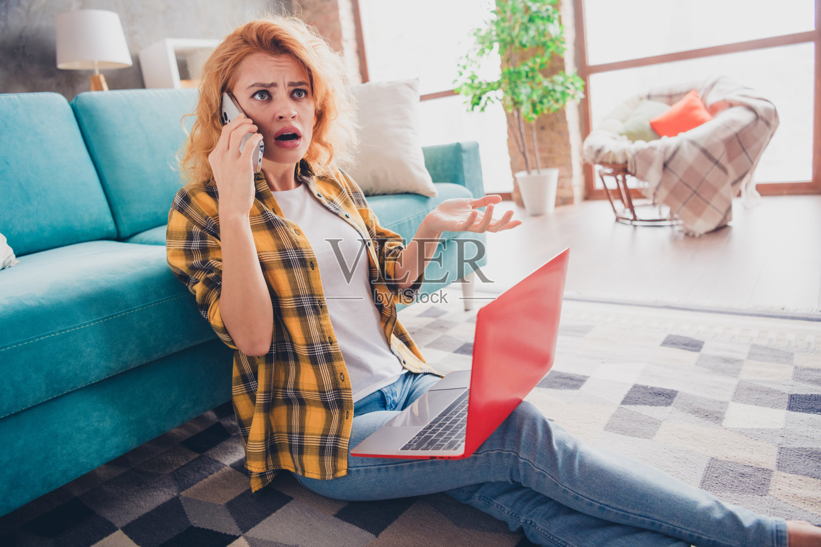 年轻金发女孩坐在地板上,穿着黄色格子衣服,使用笔记本电脑打电话,表情烦恼和害怕,身处宽敞的室内房间。照片摄影图片