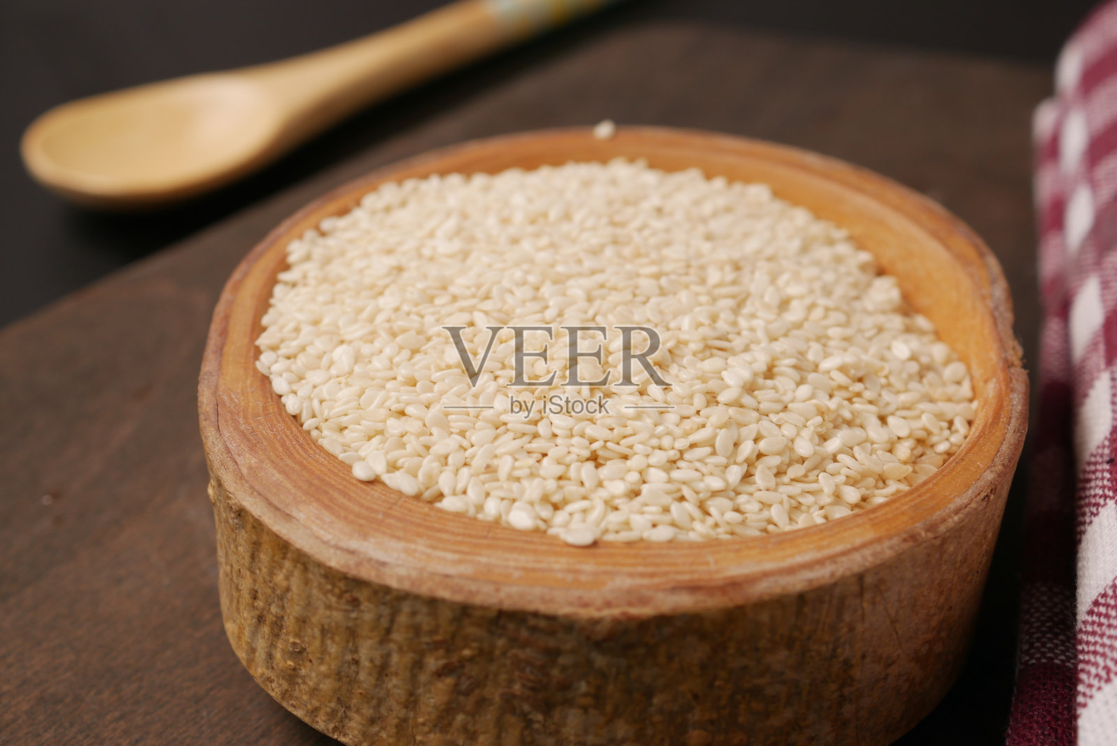 一个装满白芝麻的木碗,旁边放着一把勺子,置于深色表面上照片摄影图片