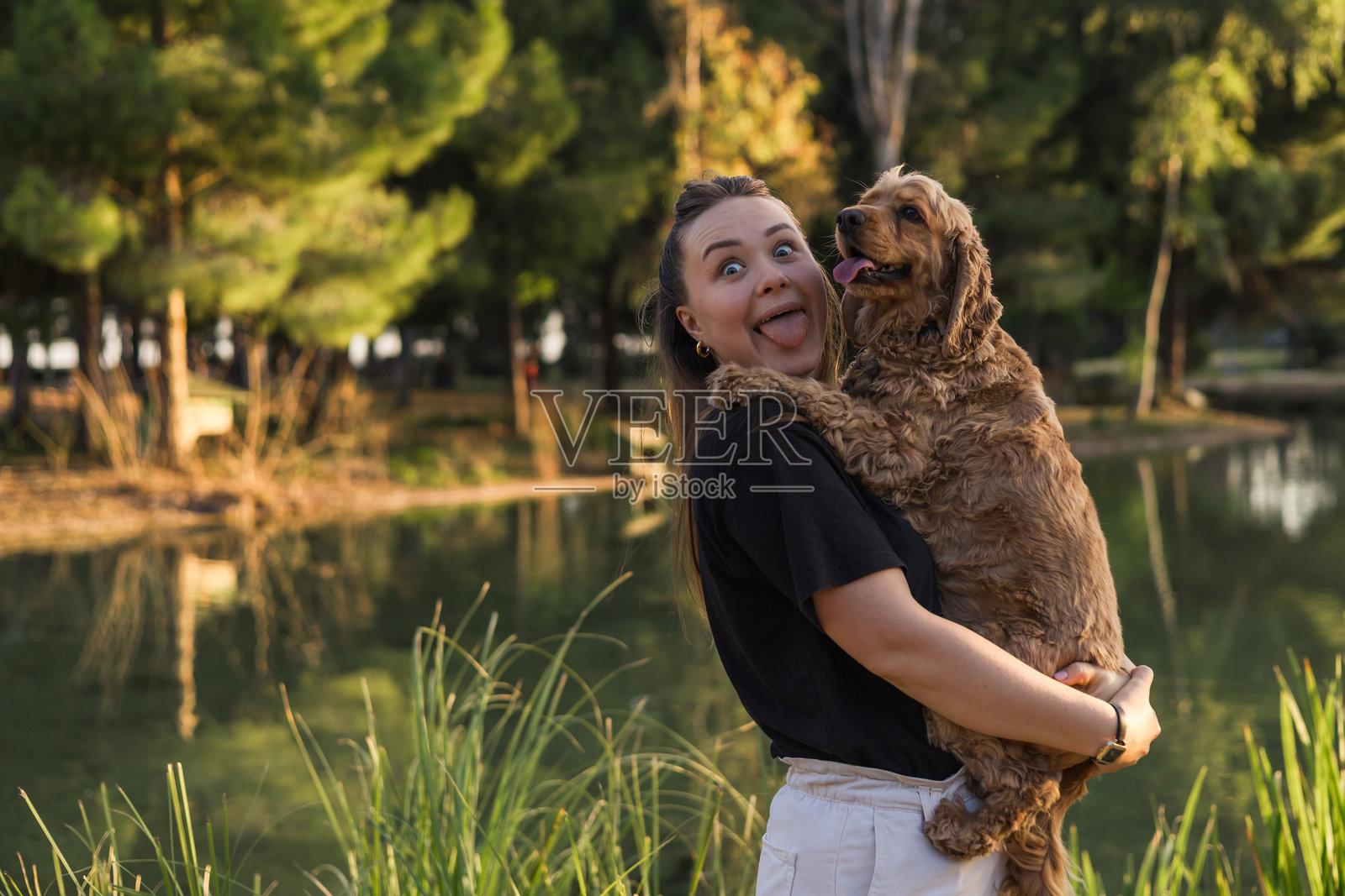 快乐的女人在宁静的公园环境中拥抱她的可卡犬,旁边是一片平静的水域。照片摄影图片