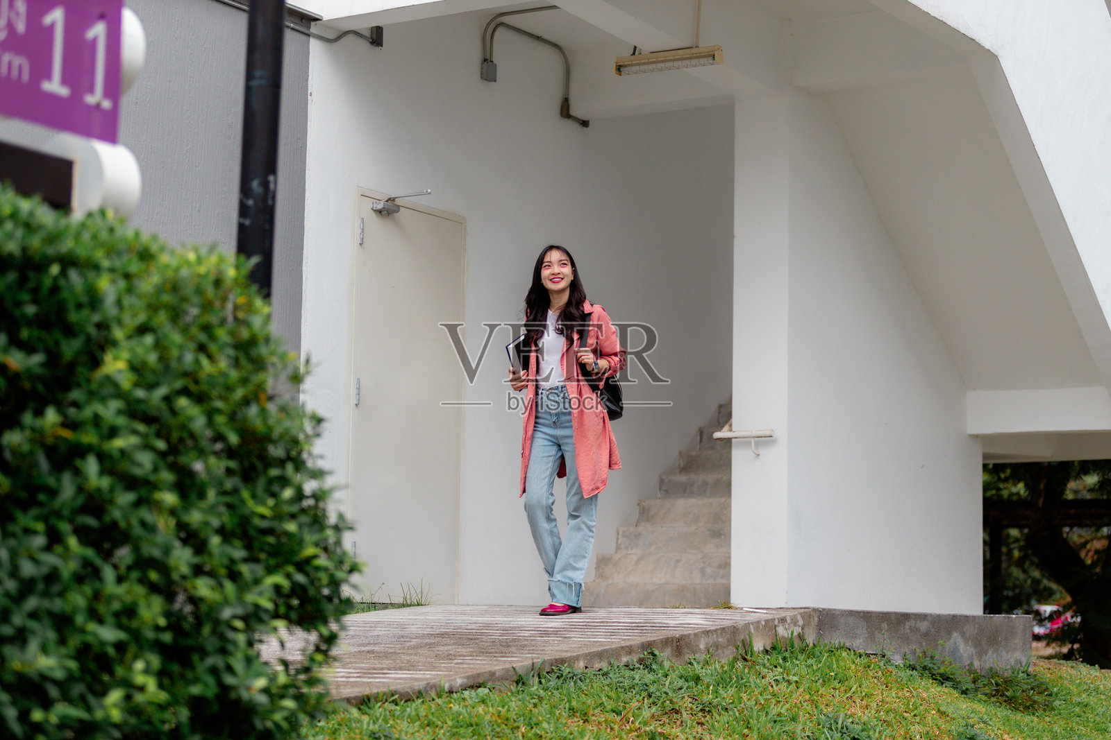 年轻的亚洲女性走出大学建筑,手里拿着书和背包,微笑着望向远方。照片摄影图片